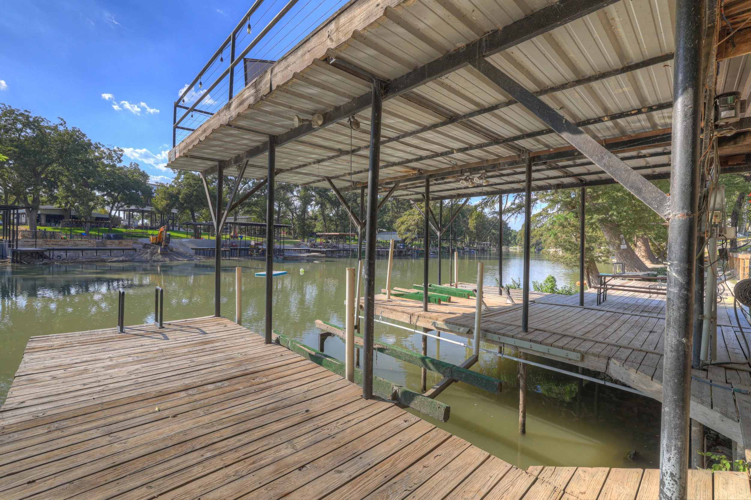 A wooden dock under construction with metal supports, overlooking a river with trees and a park in the background on a sunny day.