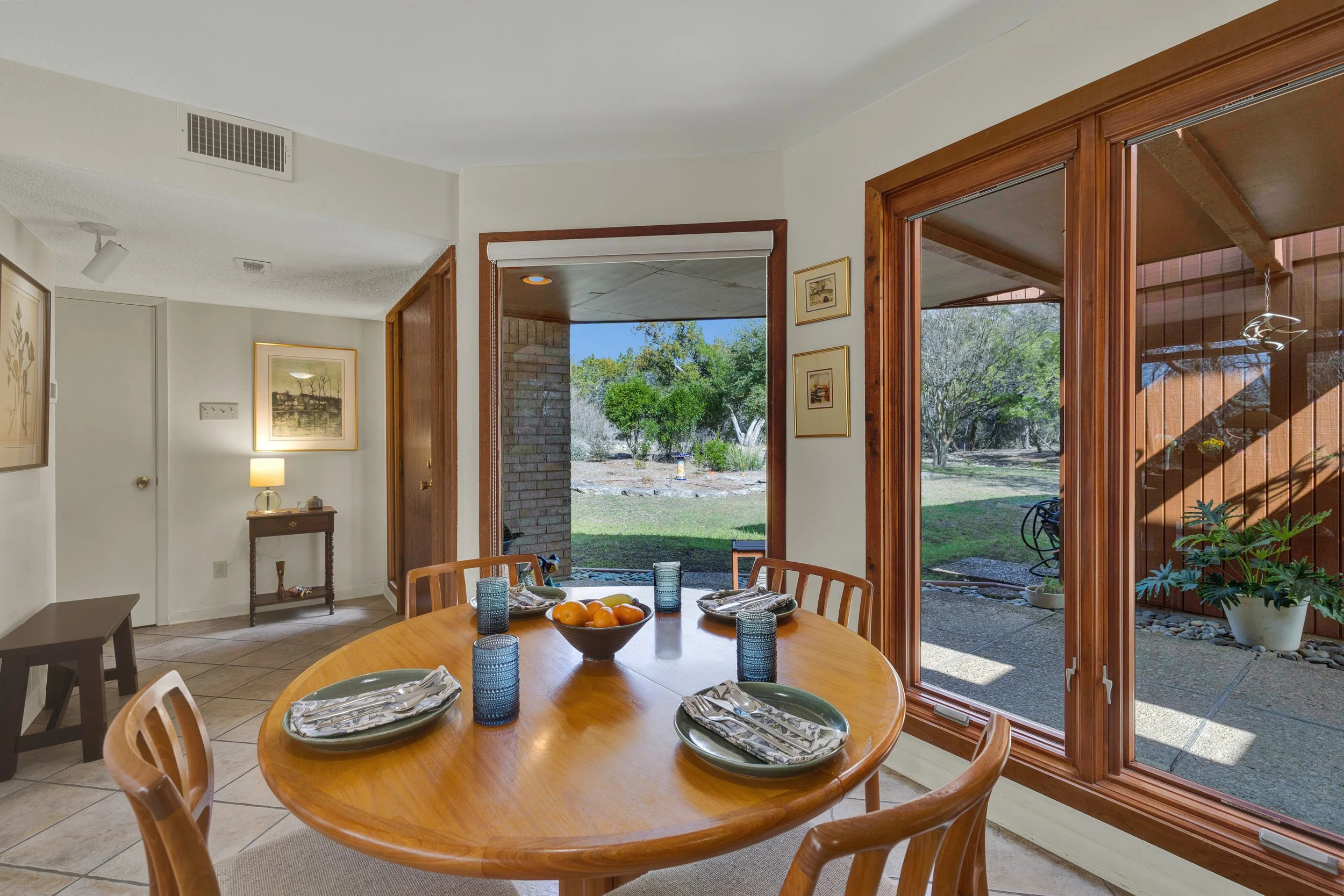 Dining room with a wooden table set with plates, glasses, and a bowl of fruit, large windows showing a backyard with greenery and trees.