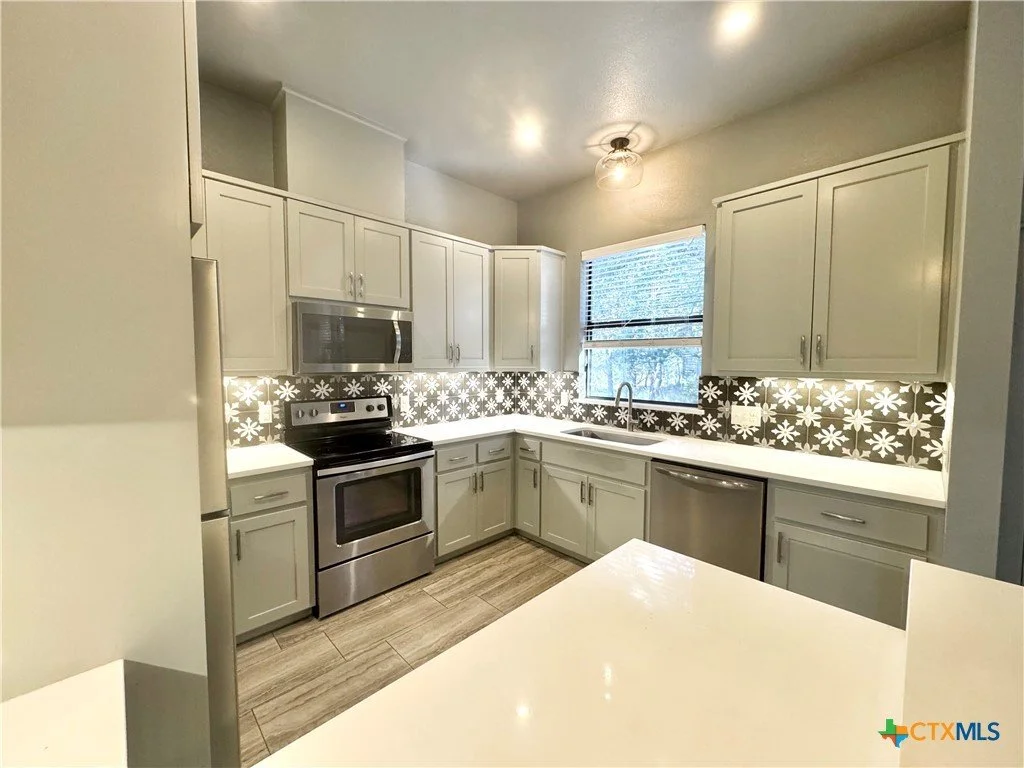 A modern kitchen with gray cabinets, stainless steel appliances, and black and white floral patterned backsplash. There is a window above the sink and a ceiling light fixture.