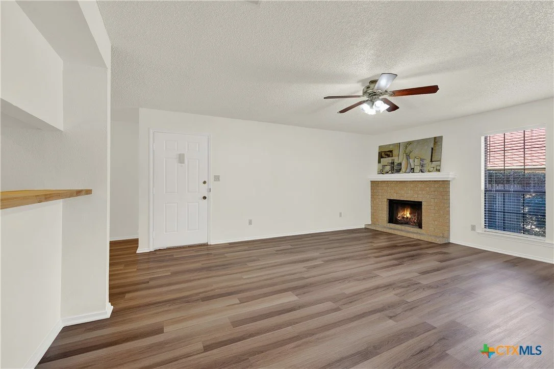 Living room with wood flooring, white walls, a fireplace with a mantel, a window with blinds, a ceiling fan, and a door.