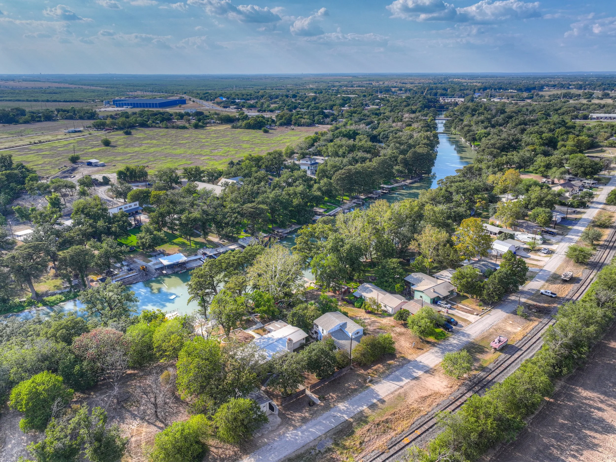 Aerial view of a suburban neighborhood with houses along a winding canal lined with trees, near railroad tracks, and open fields in the background under a partly cloudy sky.