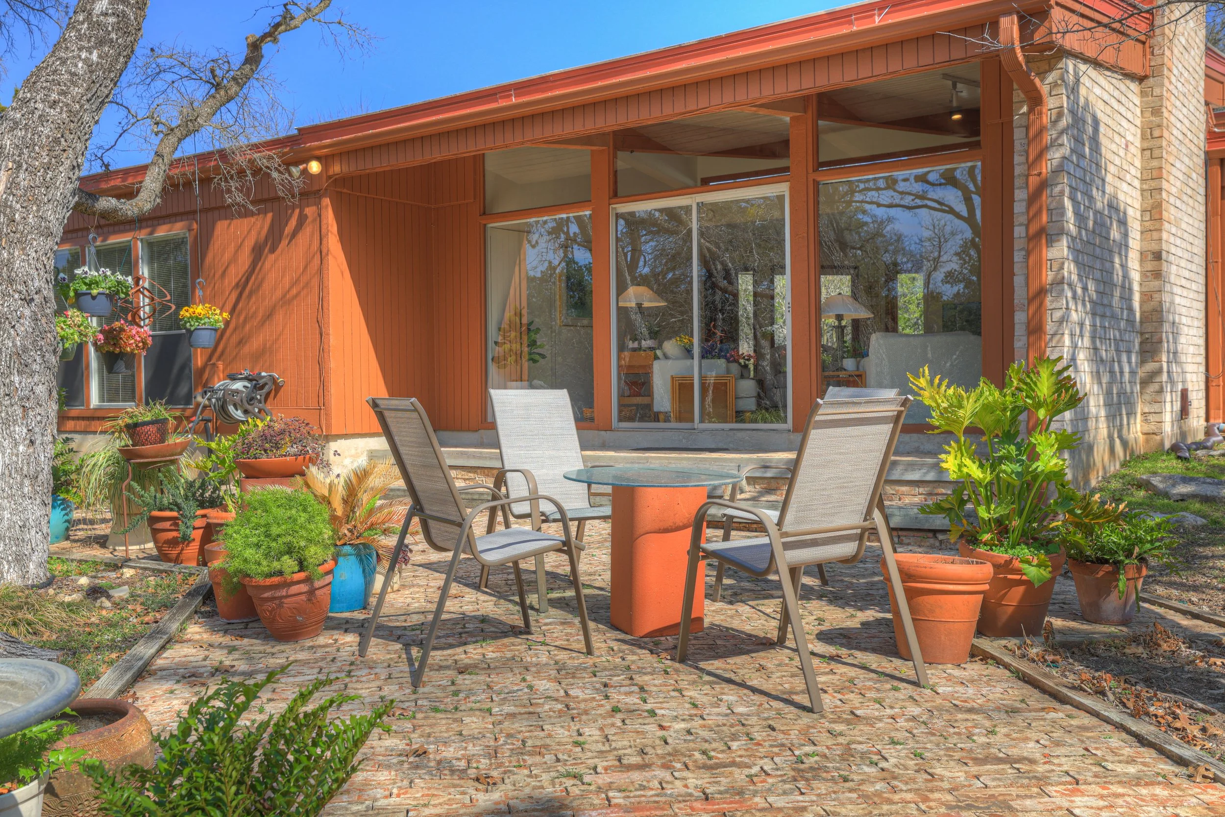 A brick patio with a round glass-top table and four outdoor chairs, surrounded by various potted plants, outside a house with large glass windows and orange siding, during daytime.