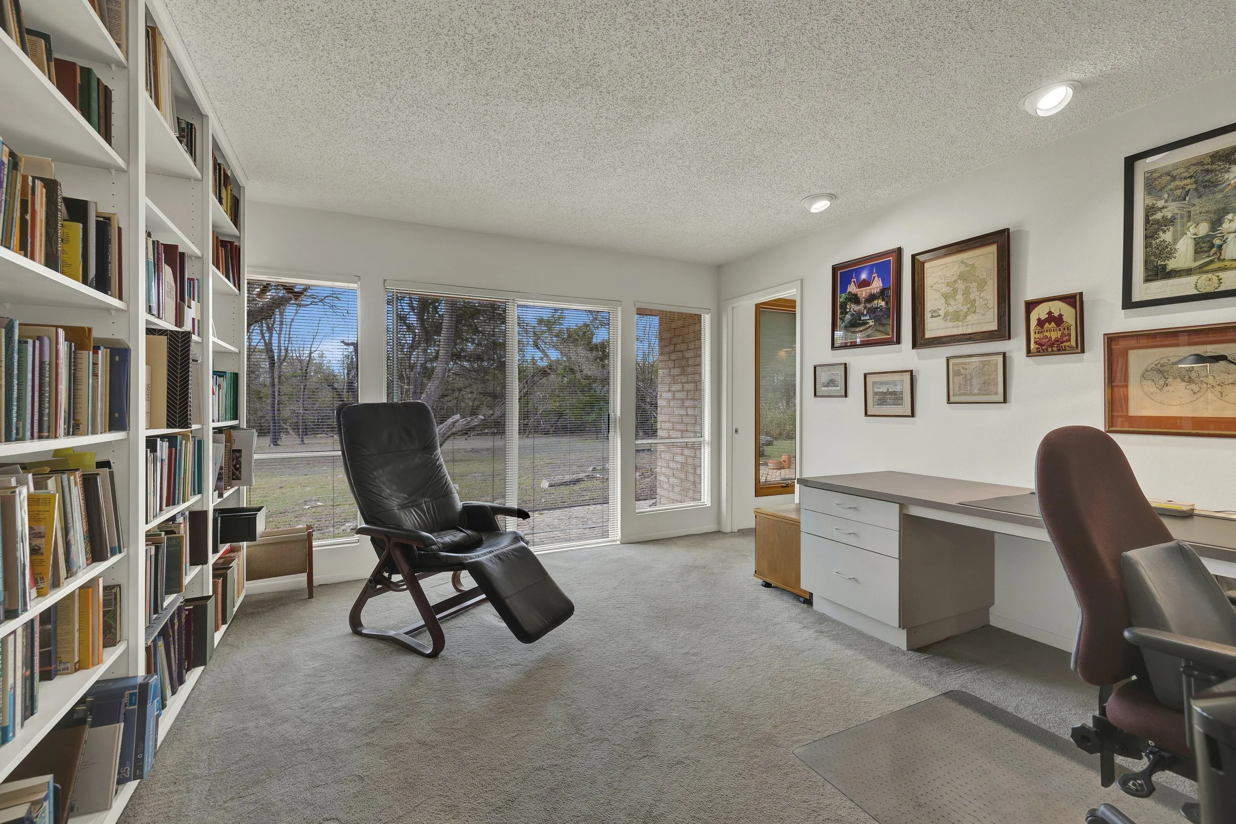 A bright home office with a large window showing an outdoor view with trees. There is a black recliner chair near the window, a white bookshelf filled with books on the left, a desk with brown and white chairs, and framed artwork on the wall.
