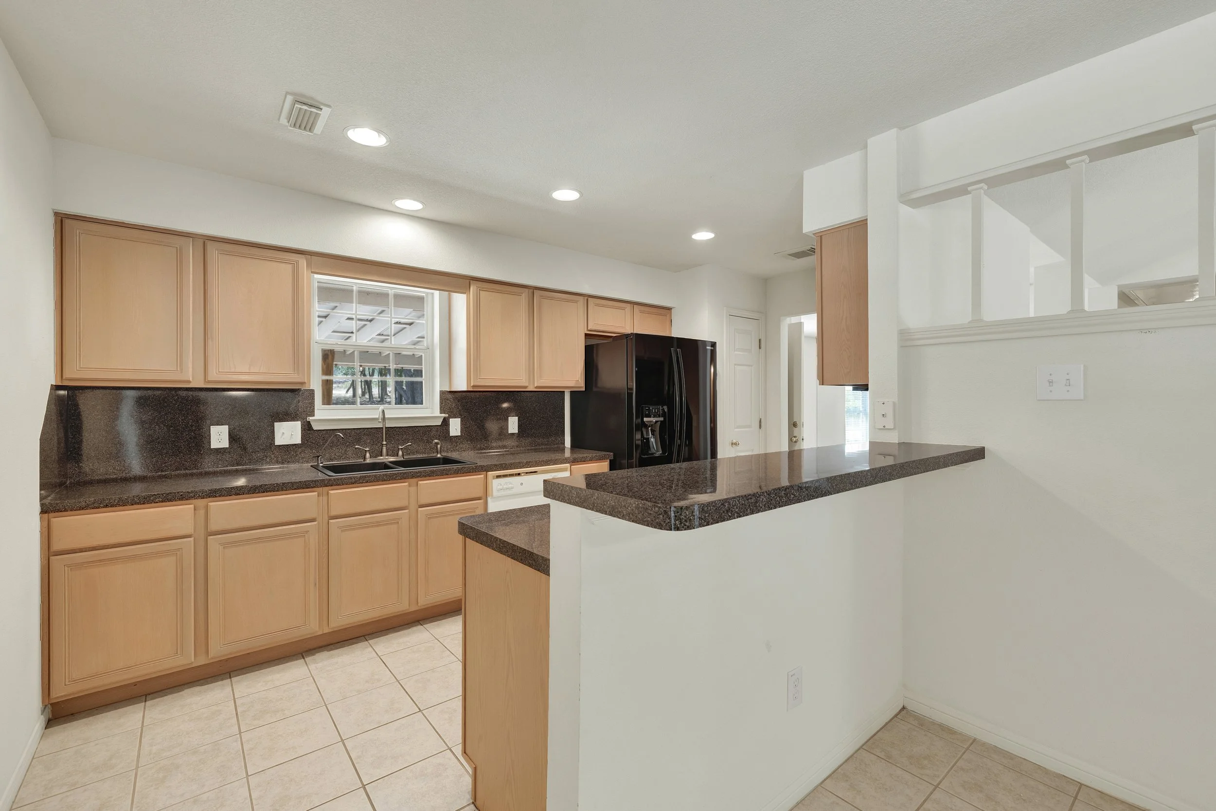 Kitchen with light wood cabinets, black refrigerator, black granite countertops, white walls, tiled floor, window above the sink, and a breakfast bar.
