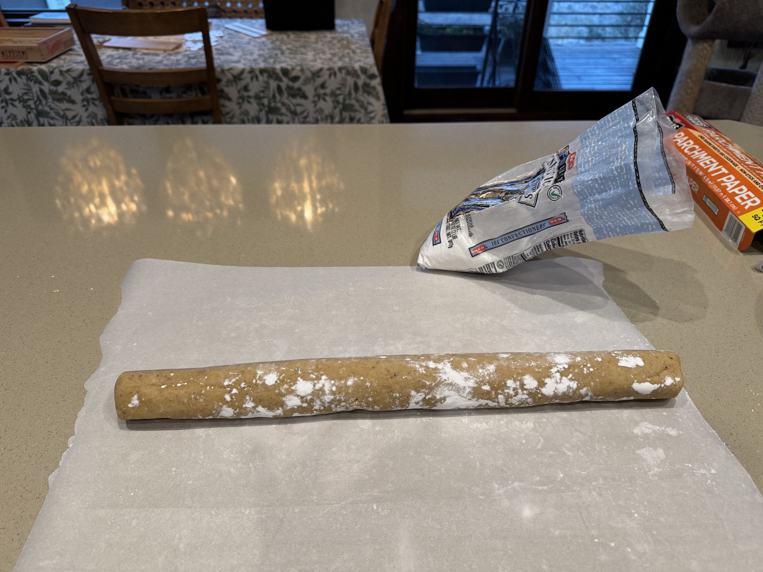 Unrolled edible cookie dough on parchment paper with a rolled cookie dough log, a partially opened bag of cookie dough, and a box of parchment paper on a kitchen counter.
