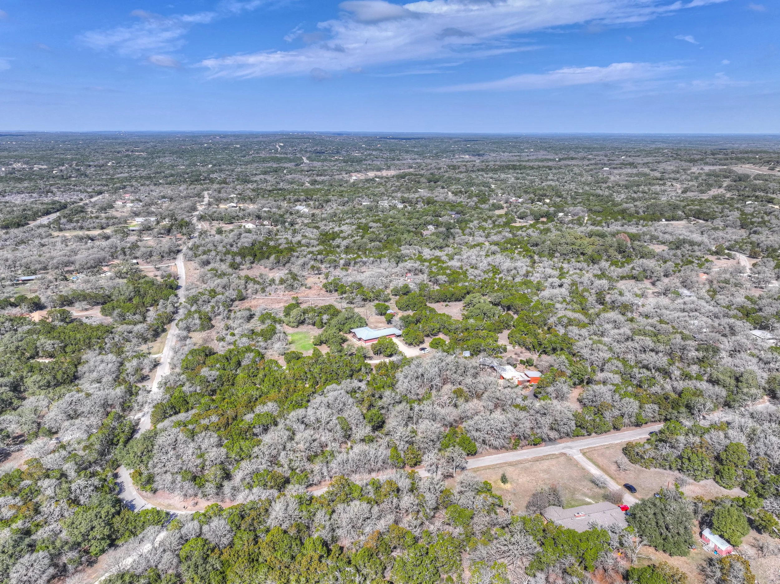 Aerial view of a rural area with densely wooded land, scattered houses, and roads under a partly cloudy sky.