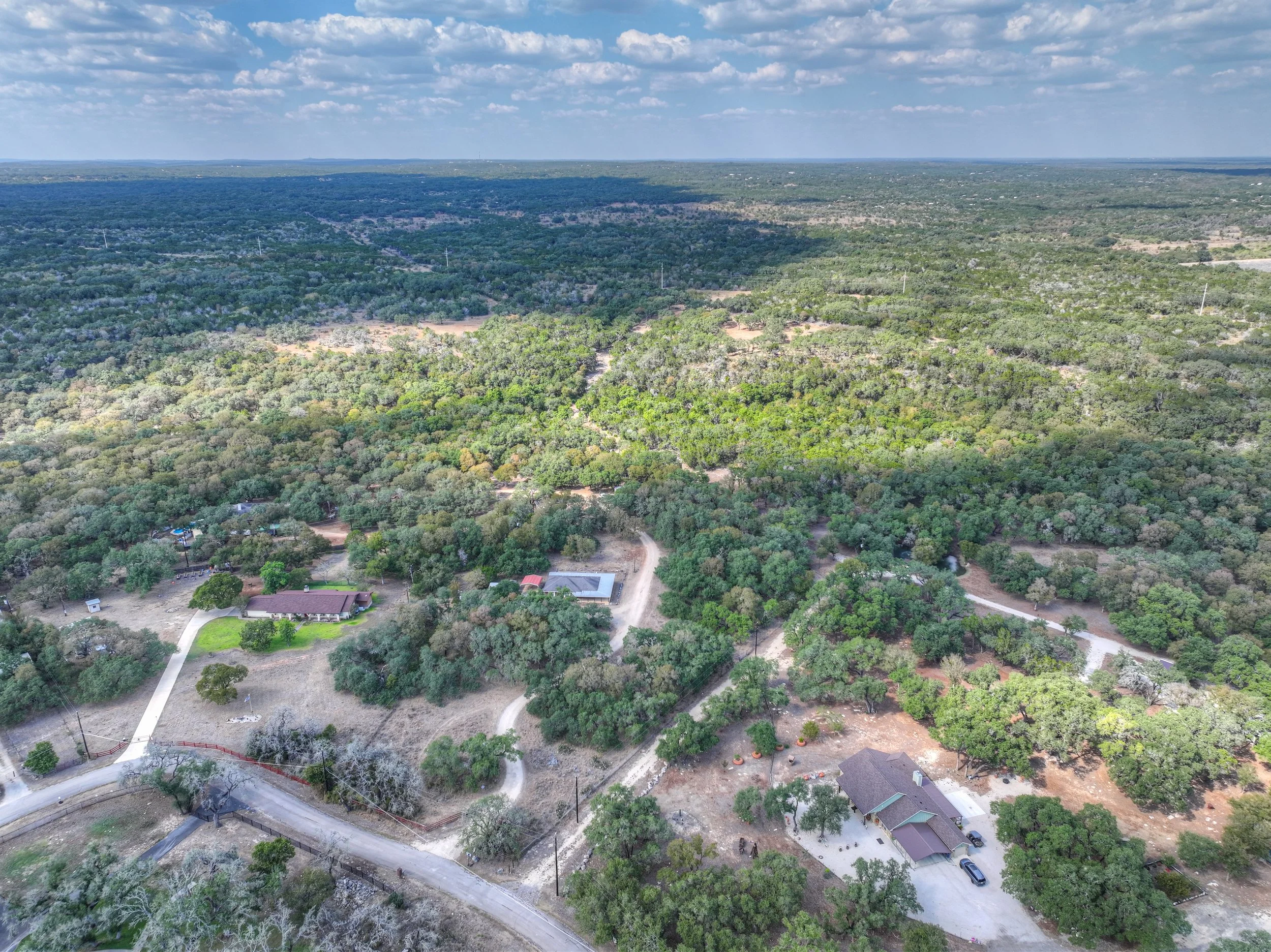 An aerial view of a suburban area surrounded by dense trees, with scattered houses, roads, and some open land under a partly cloudy sky.