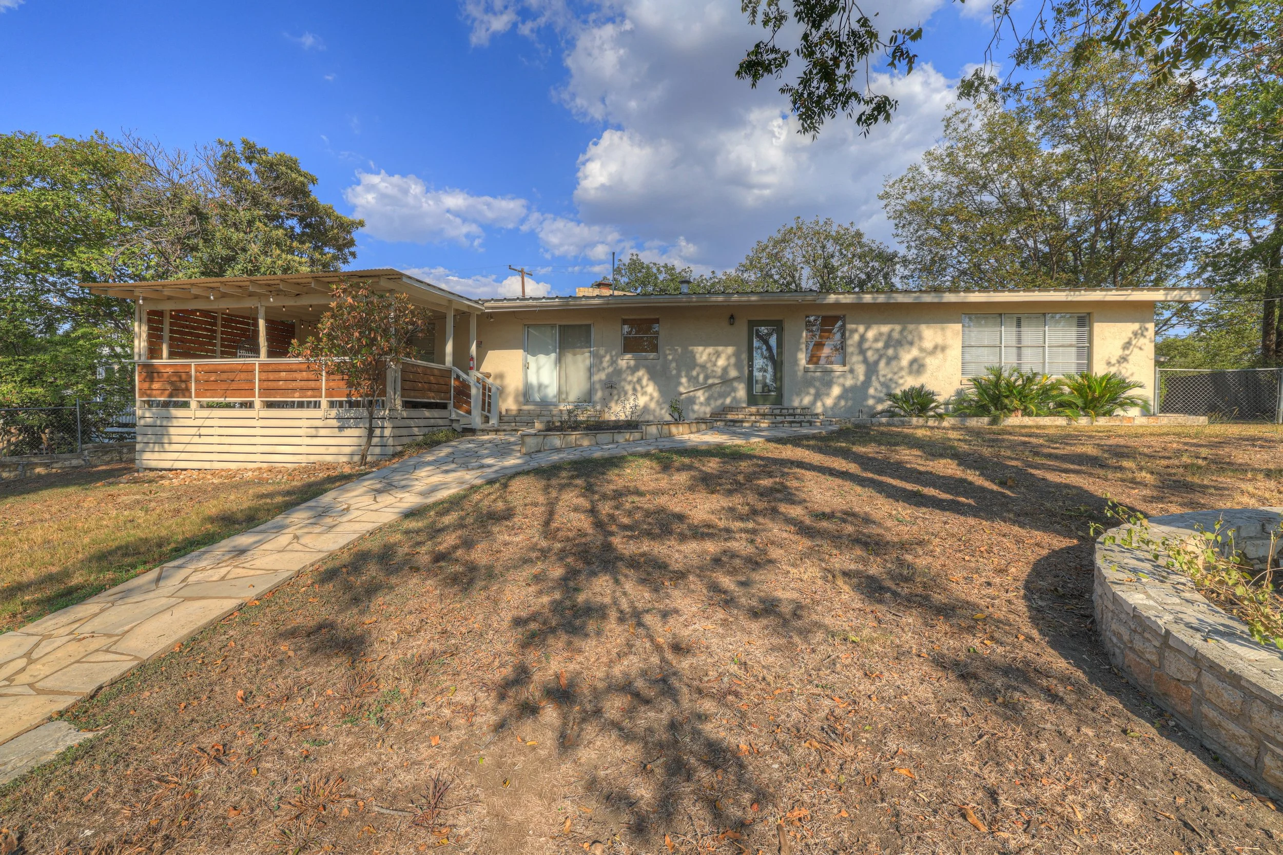 Front view of a single-story house with a stone pathway leading up to the porch, beige exterior walls, and varied windows. The house features a raised porch with a wooden railing and several plants and trees surrounding it. The yard has dry grass and