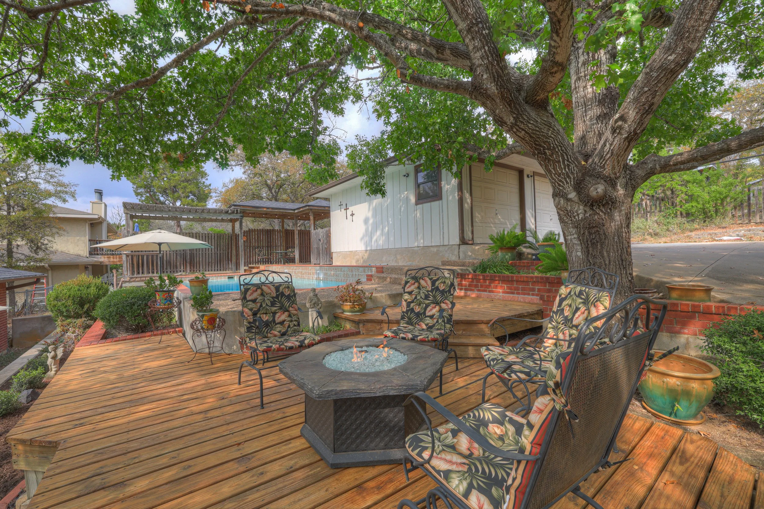 A backyard patio with a wooden deck, patio chairs with floral cushions around a fire pit, a large tree providing shade, potted plants, and a pool with an umbrella nearby.