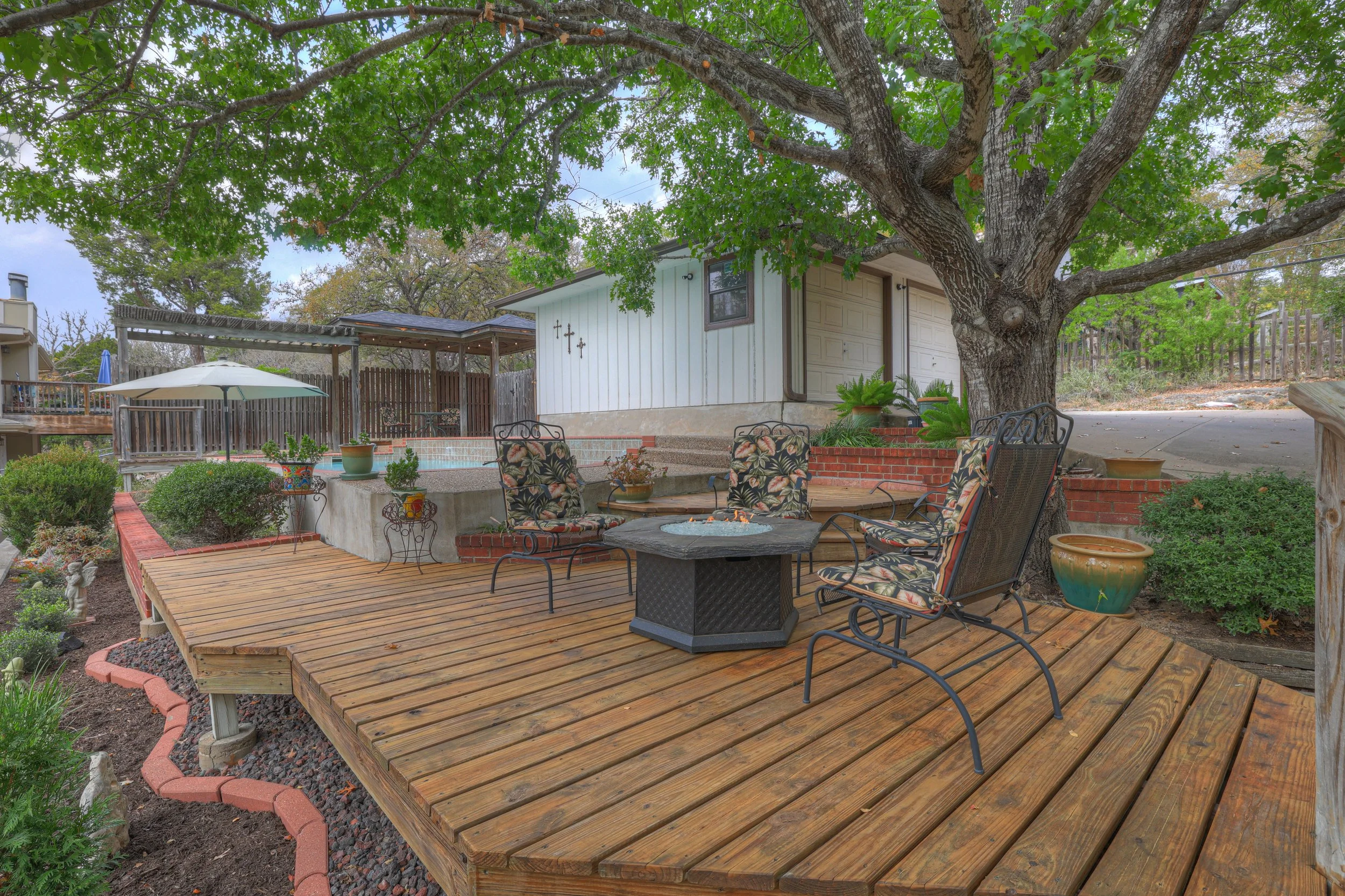 Backyard patio with a large tree, wooden deck, outdoor chairs with floral cushions, small fire pit, potted plants, and a hot tub, with a white house in the background.