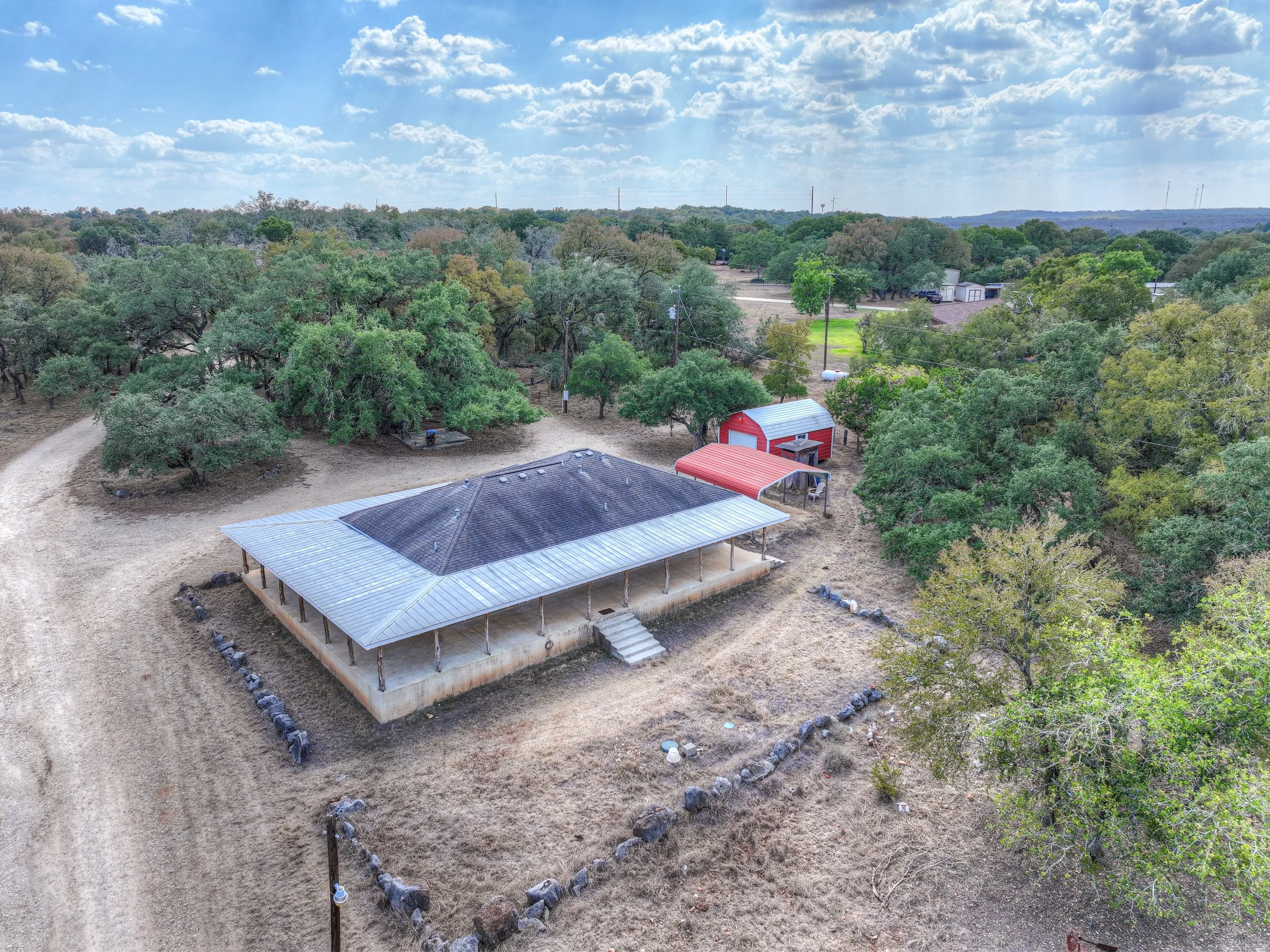 An aerial view of a rural property featuring a house with a metal roof, a red outbuilding, and trees surrounding the area on a sunny day with clear skies.