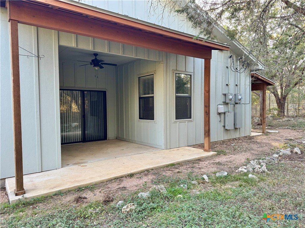 Back porch area of a house with a concrete slab, sliding glass door, two windows, a ceiling fan, and outdoor utility meters on the wall. There's a wooden support beam and a sloped roof over the porch.