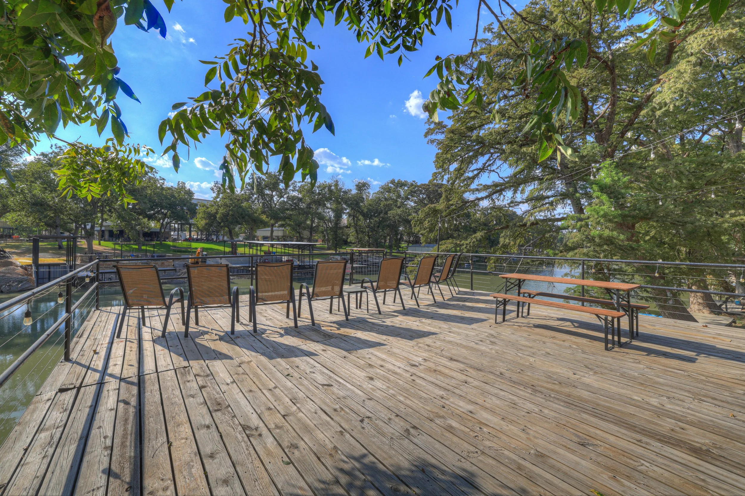 A spacious wooden deck with metal railings overlooking a pond, with several brown chairs and two benches arranged on the deck. The surrounding area has lush green trees, a bright blue sky with some clouds, and a grassy lawn in the background.
