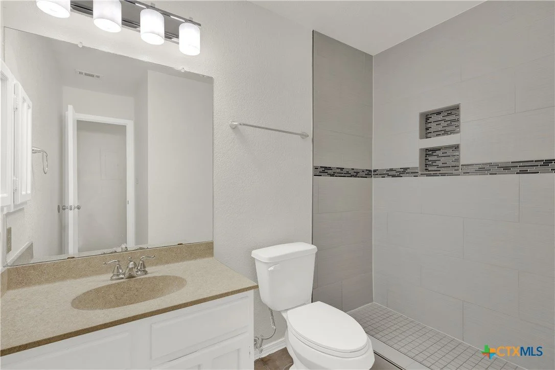 Bathroom with a white vanity, beige countertop, large mirror, white toilet, and a walk-in shower with gray tiles and built-in shelves.