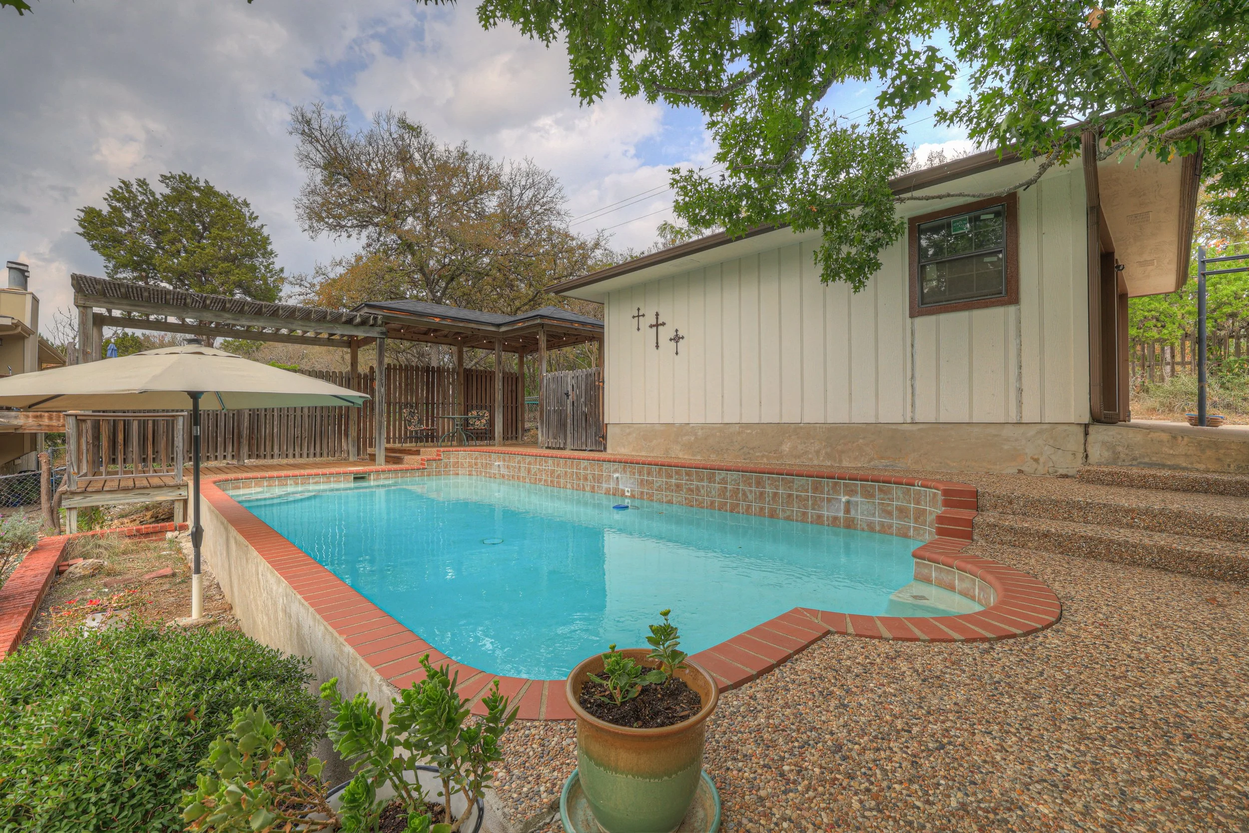 Backyard with a swimming pool, umbrella, potted plant, and a small structure with religious crosses on the wall.