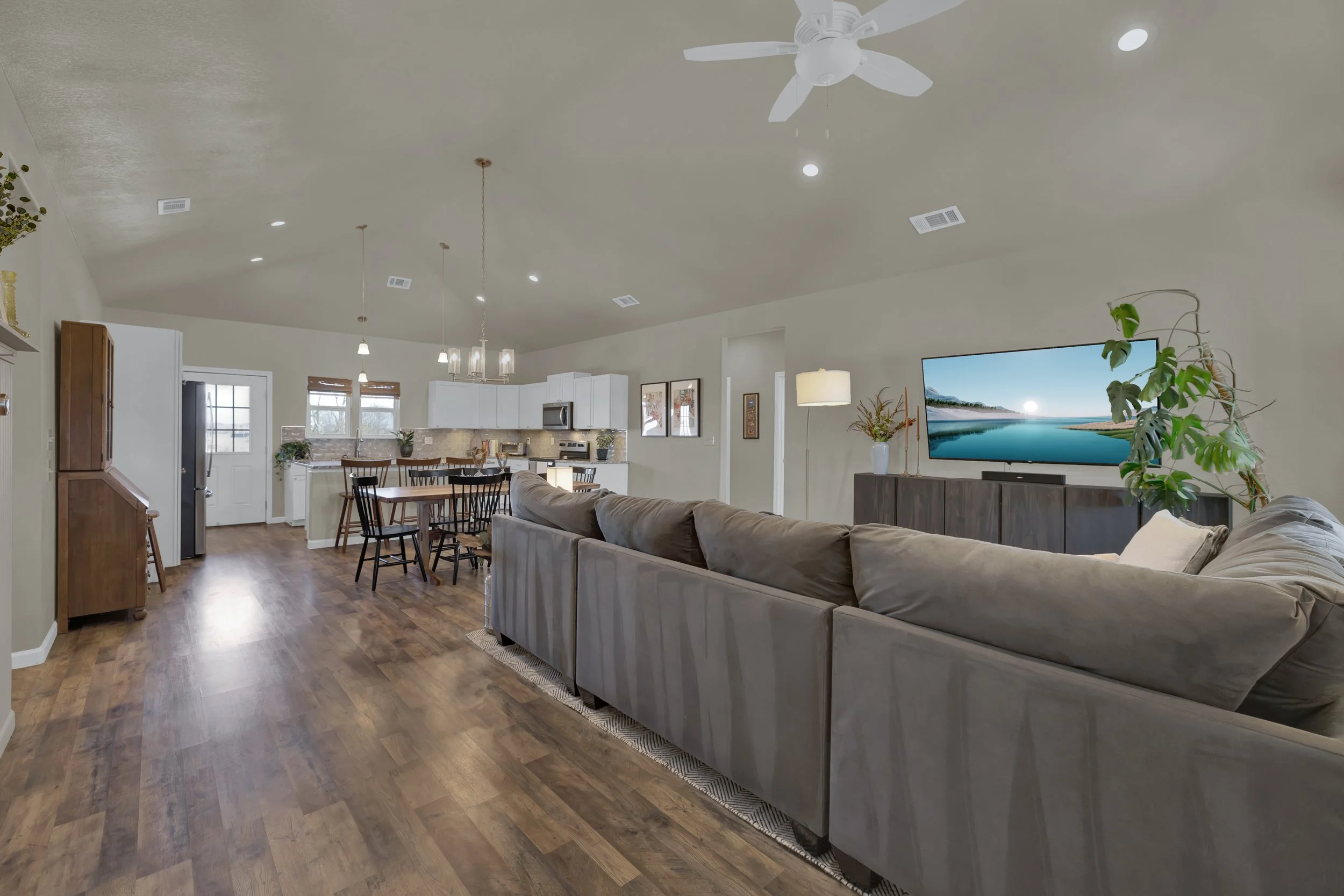 Open-concept living room and kitchen with hardwood floors, gray sectional sofa, flat-screen TV, white kitchen cabinets, and dining area with wooden table and black chairs.