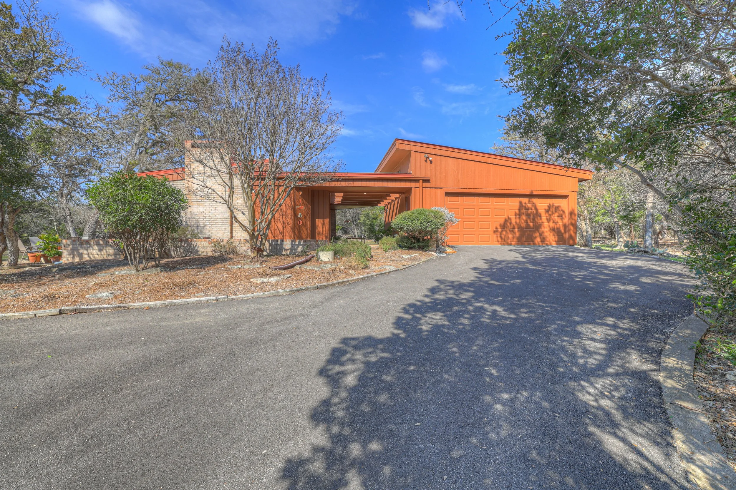 Front view of a house with a red exterior, attached garage, and a driveway with shadows from trees, under a blue sky.