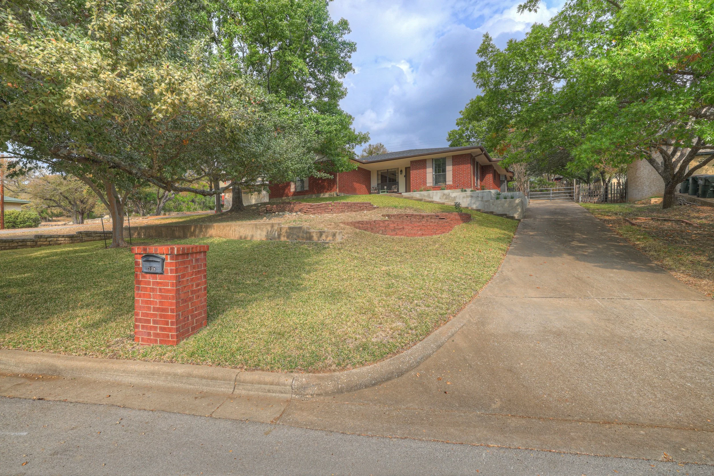 A suburban house with a brick exterior, situated on a grassy hill with large trees providing shade. A concrete driveway leads up to the house, and a brick mailbox is in the front yard.