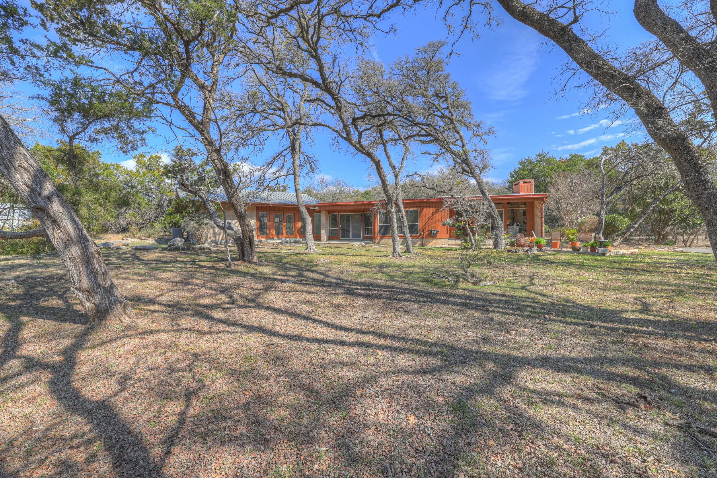 A modern single-story house with large windows and sliding glass doors, surrounded by leafless trees and a green grassy yard under a blue sky.