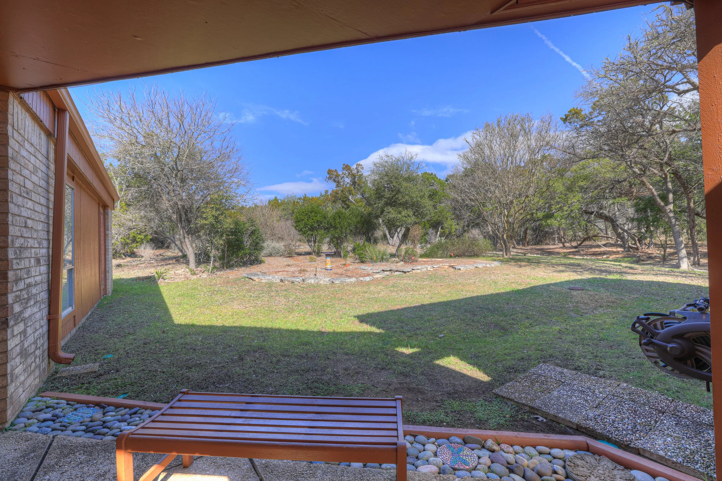 View of a backyard with a grassy lawn and several trees, seen from a covered patio area with a wooden bench, a pebble border, and a place for outdoor decorations. The sky is bright blue with a few clouds and a visible contrail.