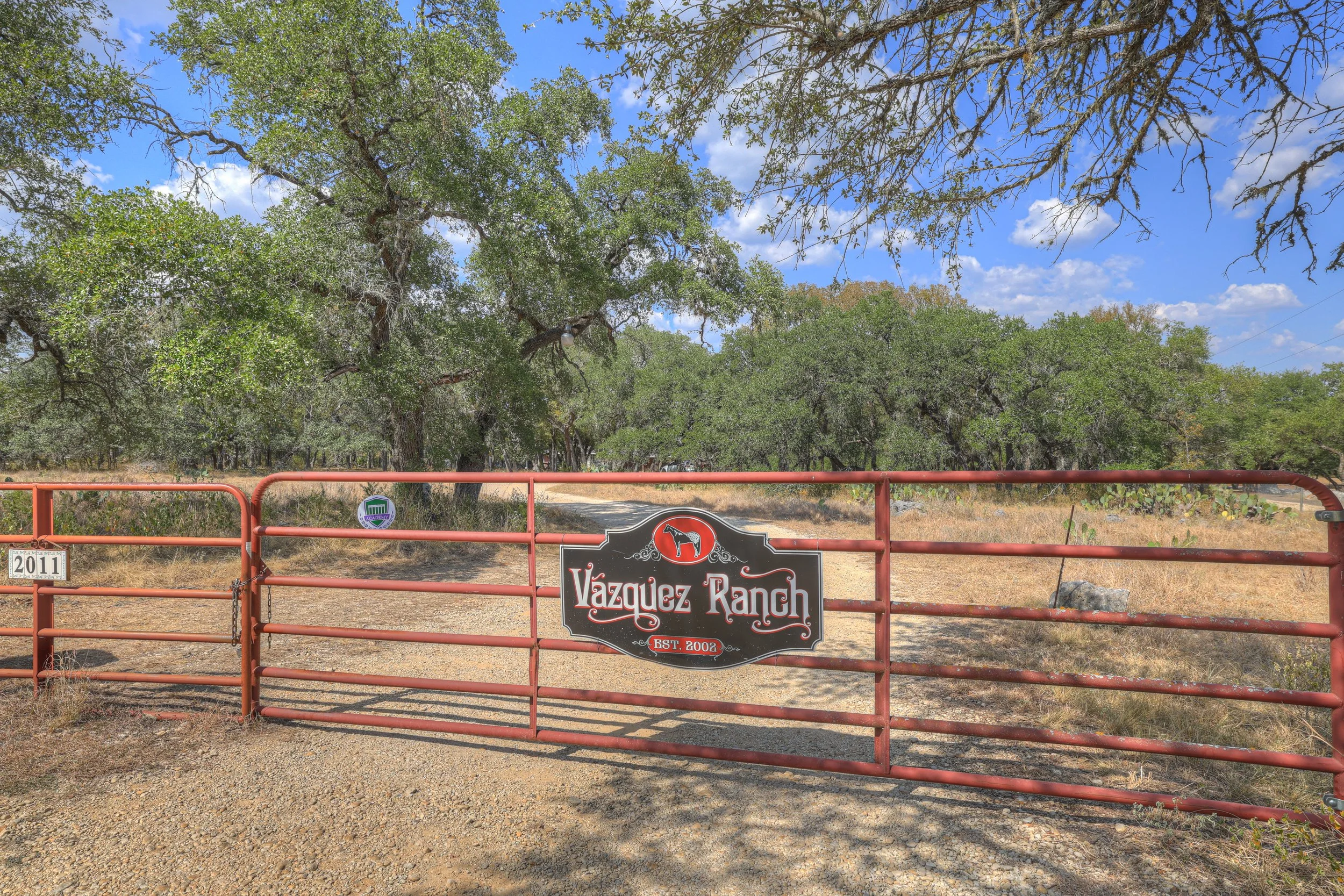 A red metal gate with a sign reading 'Vázquez Ranch' and established in 2002, set in a rural landscape with trees, blue sky, and a dirt road beyond.