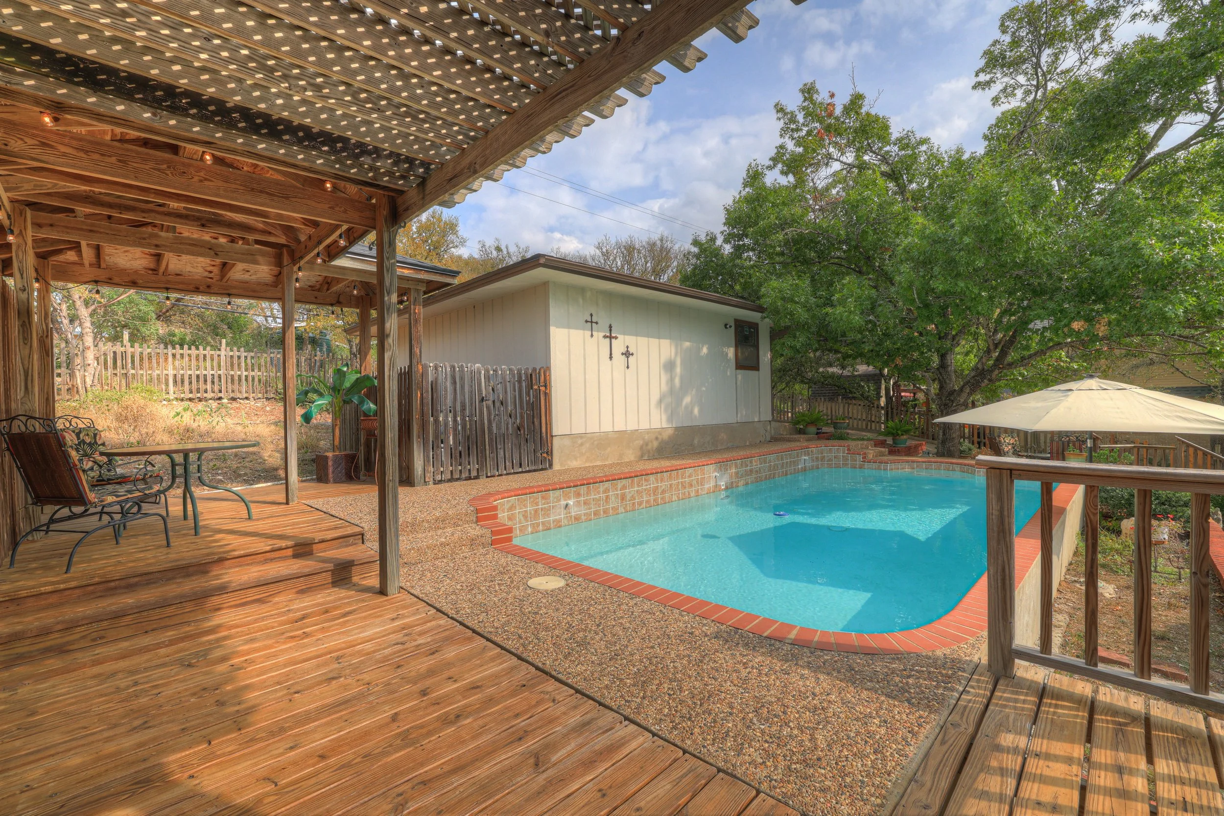 Backyard swimming pool with a wooden deck and shaded patio area, surrounded by a wooden fence, trees, and outdoor furniture.