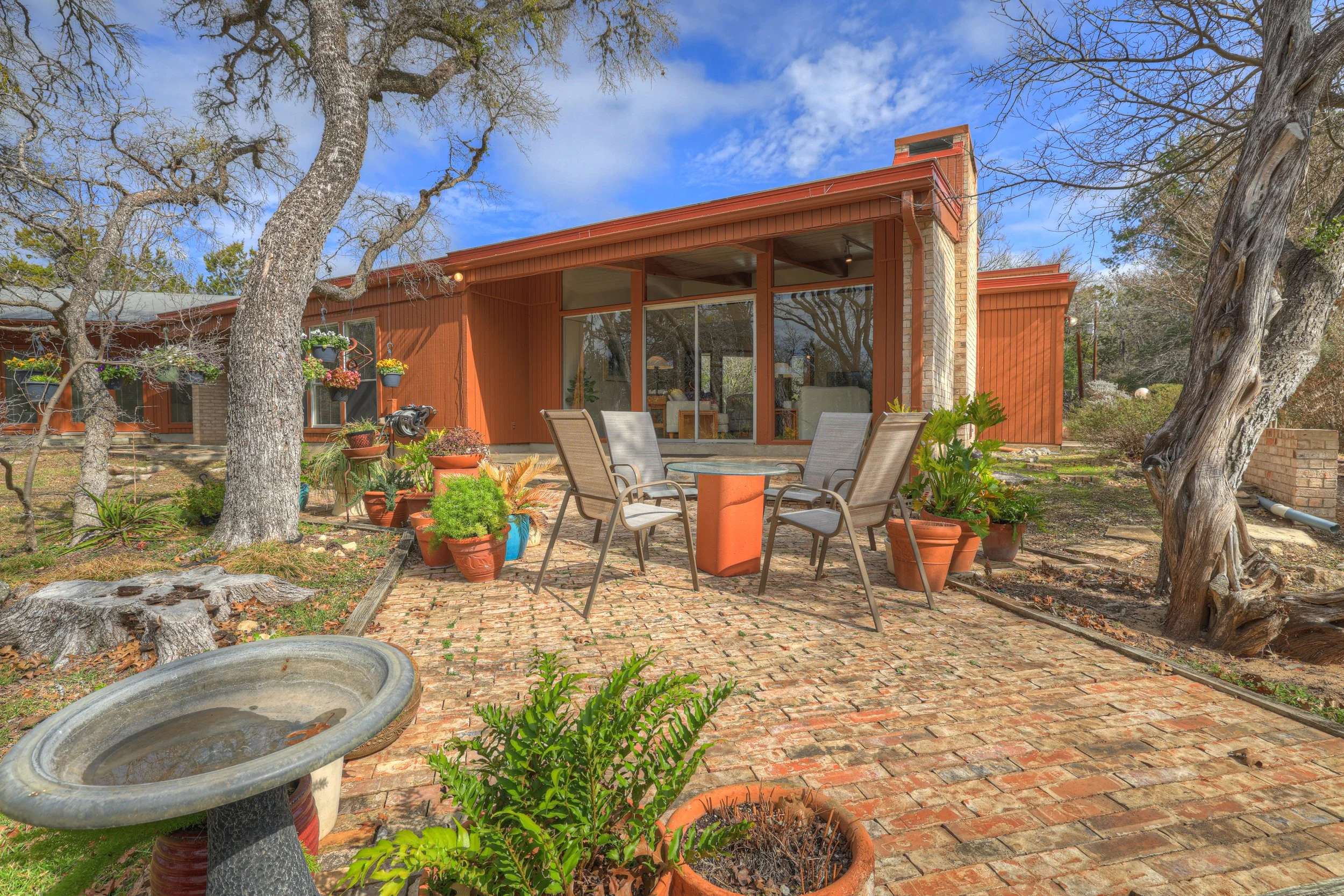 A cozy backyard patio with a brick floor, surrounded by potted plants, trees, and a wooden house with large glass sliding doors under a partly cloudy sky.
