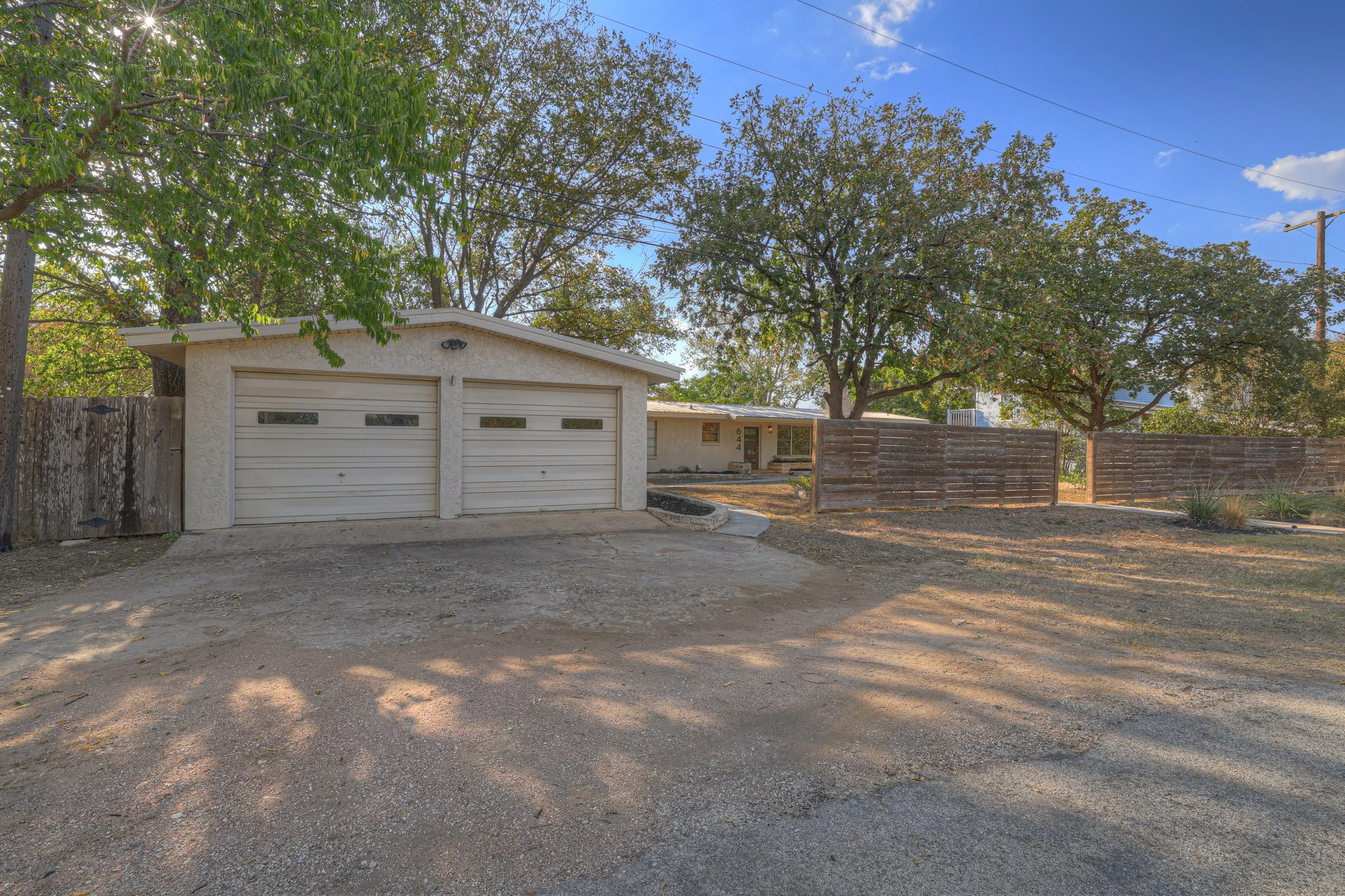 Front yard of a single-story house with a two-car garage, trees, and a wooden fence on a sunny day.