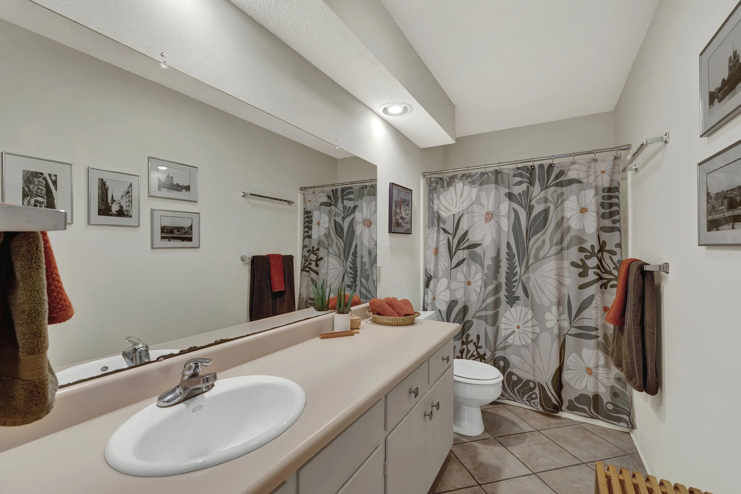 Bathroom with a large mirror, beige countertop with sink, patterned shower curtain, framed black-and-white photos on the wall, and towels hanging on the wall.