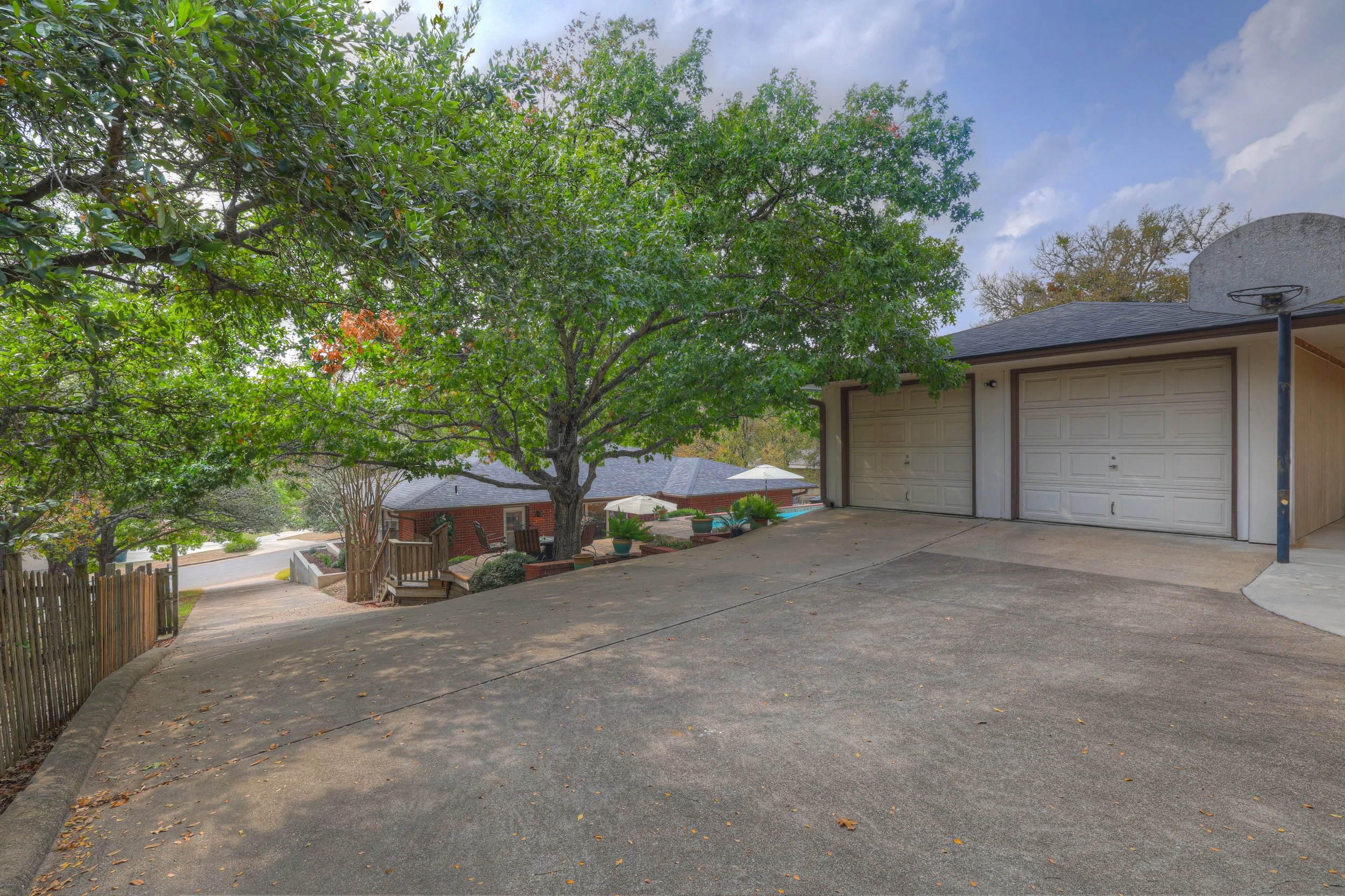 Residential driveway with a double garage, large tree, potted plants, wooden staircase, and a basketball hoop, in a suburban neighborhood.