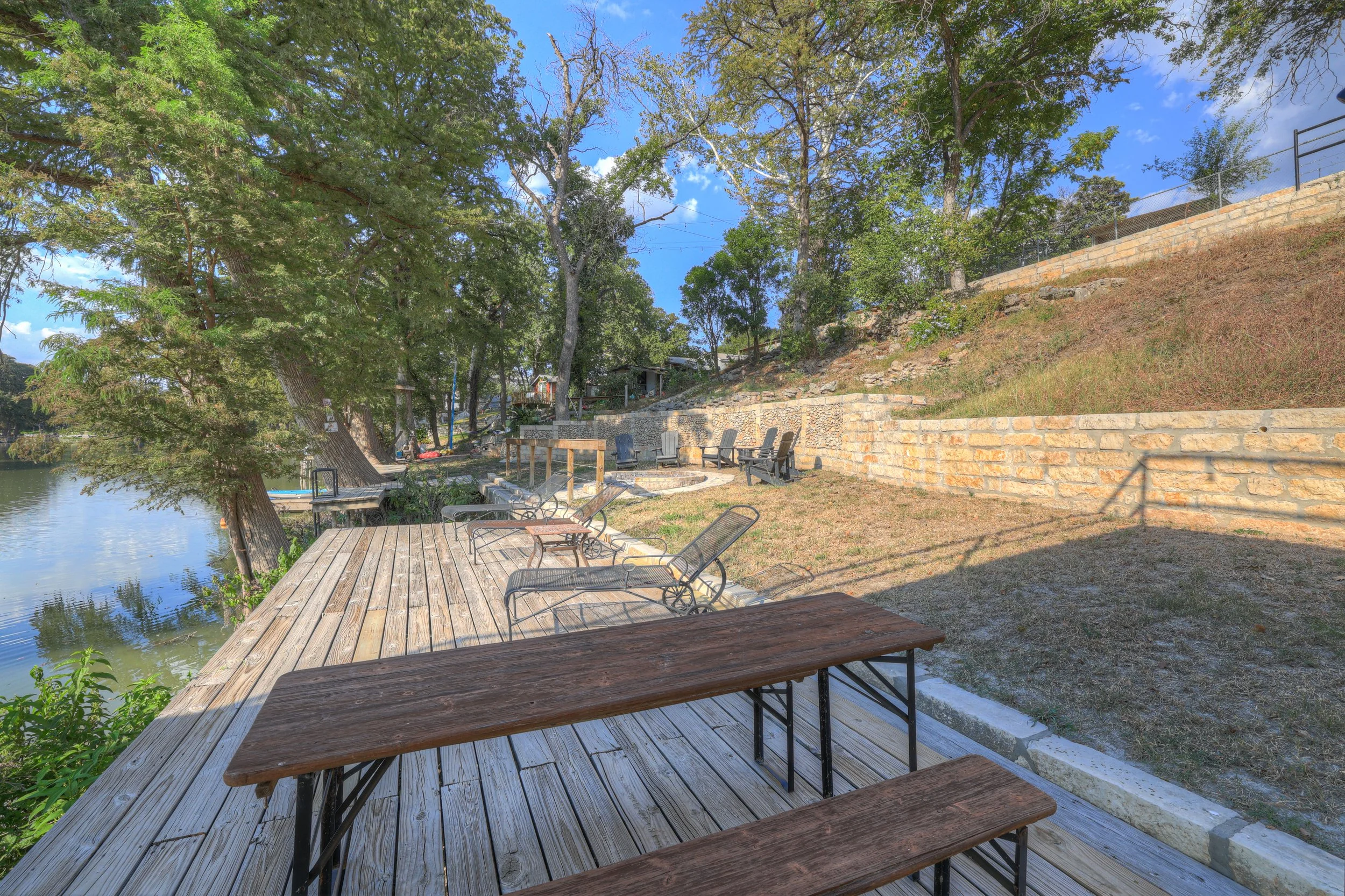 View of a riverside outdoor seating area with wooden deck, various chairs, trees, and a stone retaining wall under a partly cloudy sky.
