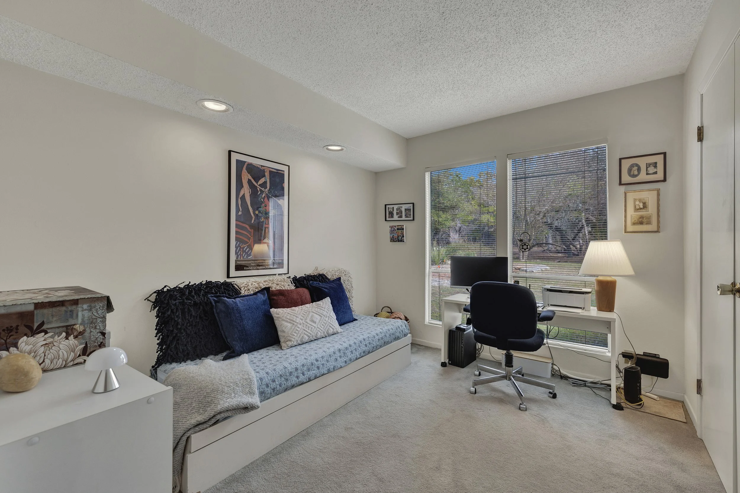A cozy living room with a daybed decorated with dark blue and white pillows, a desk with a computer and a lamp, a window with blinds showing trees outside, and framed pictures on the wall.