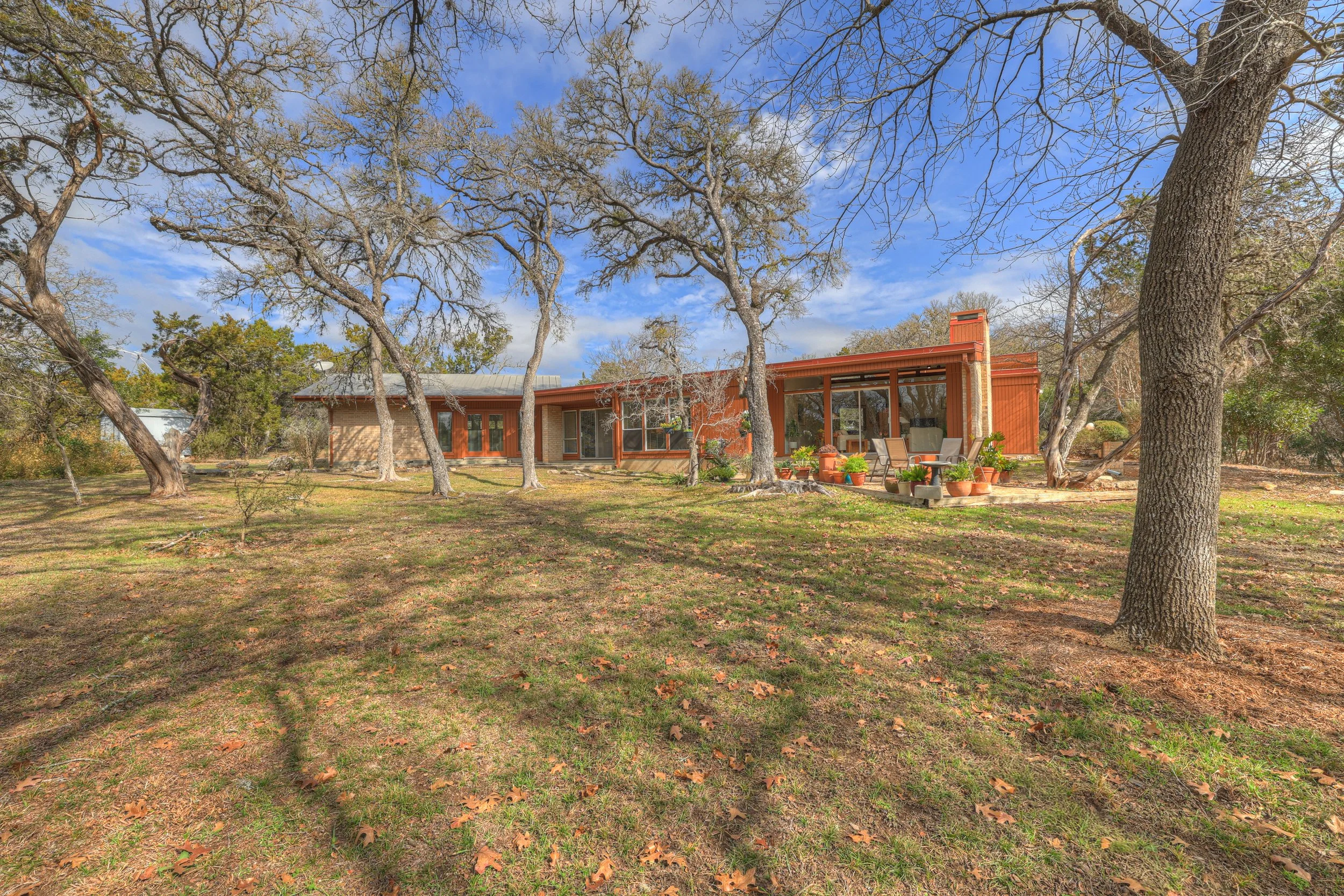 A mid-century style house with large glass windows and a patio with outdoor chairs and potted plants, surrounded by deciduous trees on a grassy yard.