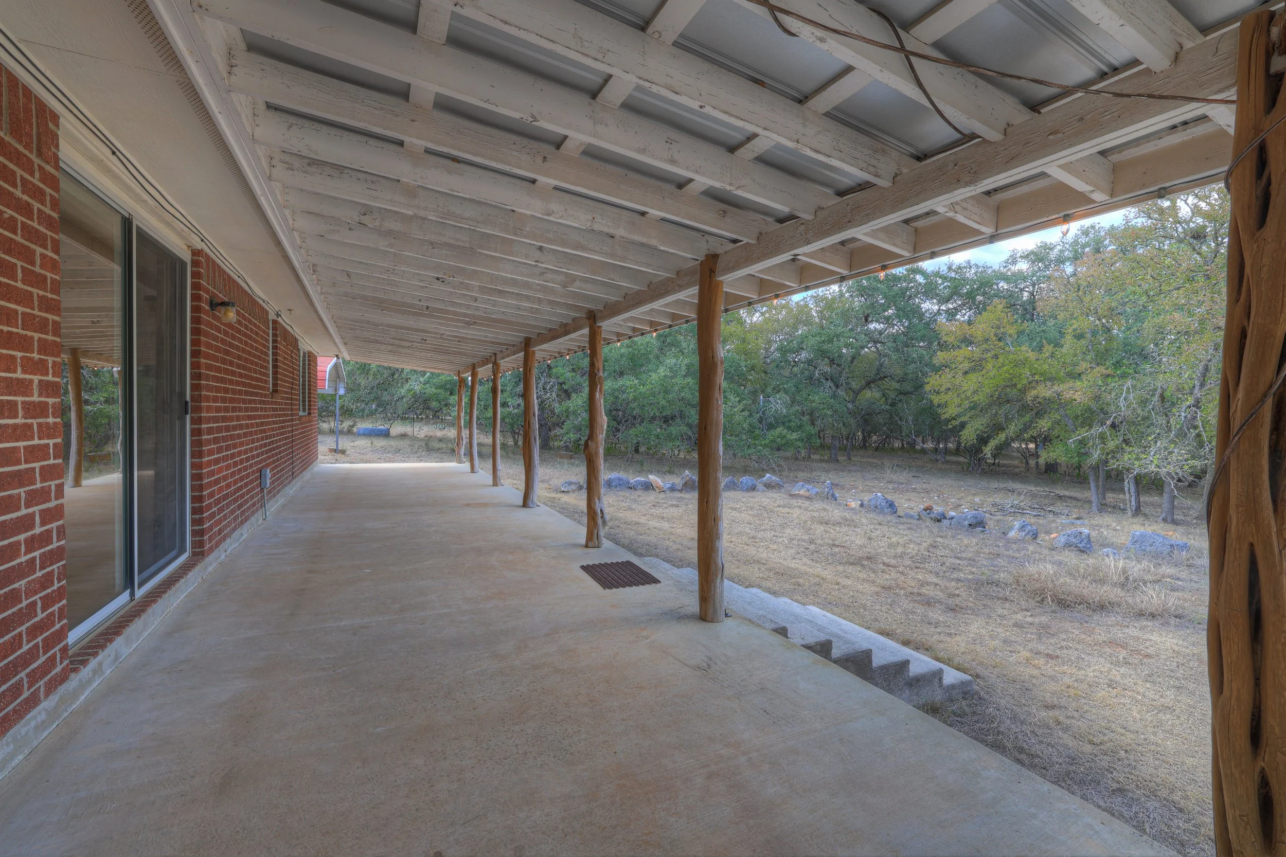 Covered porch with wooden support beams, a concrete floor, brick wall with sliding glass door, and view of a yard with grass, rocks, and trees.