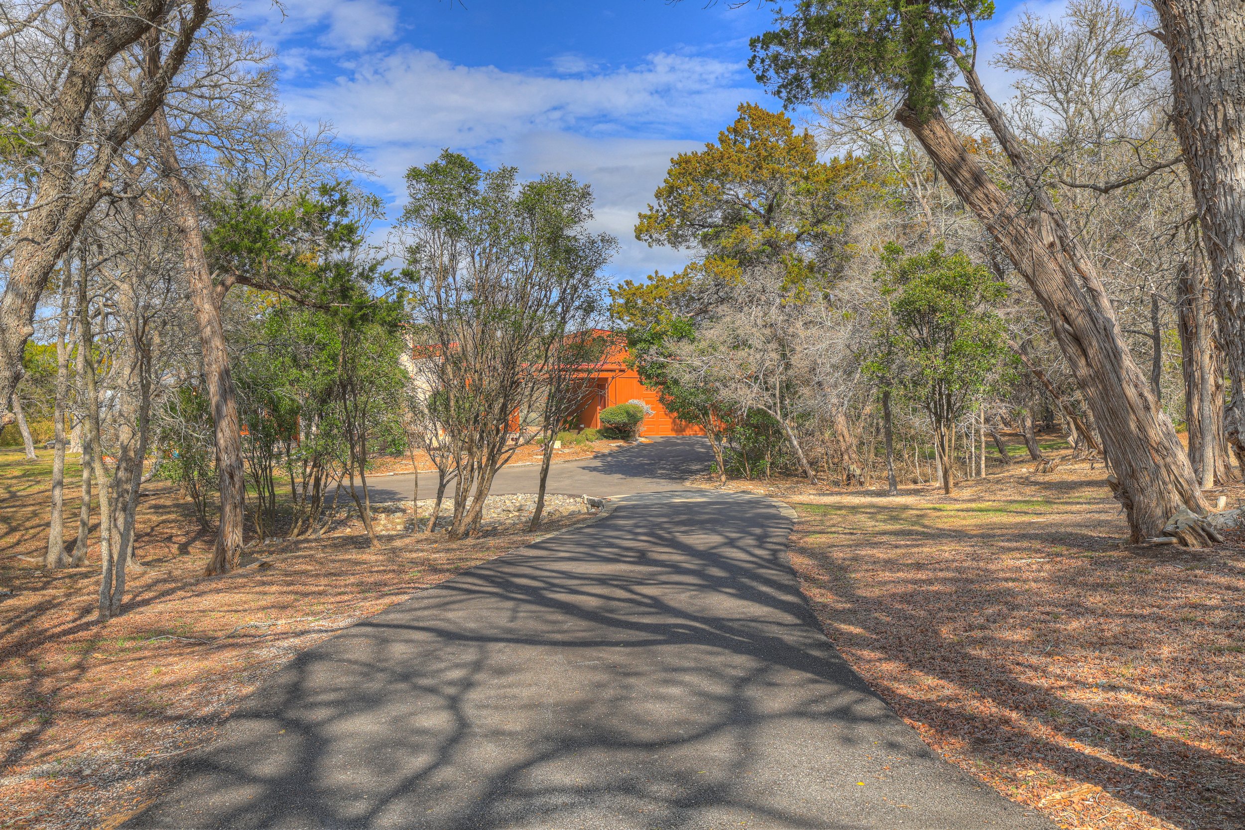 A winding paved driveway leads to a building partially obscured by trees, with leafy and bare branches, under a partly cloudy blue sky.