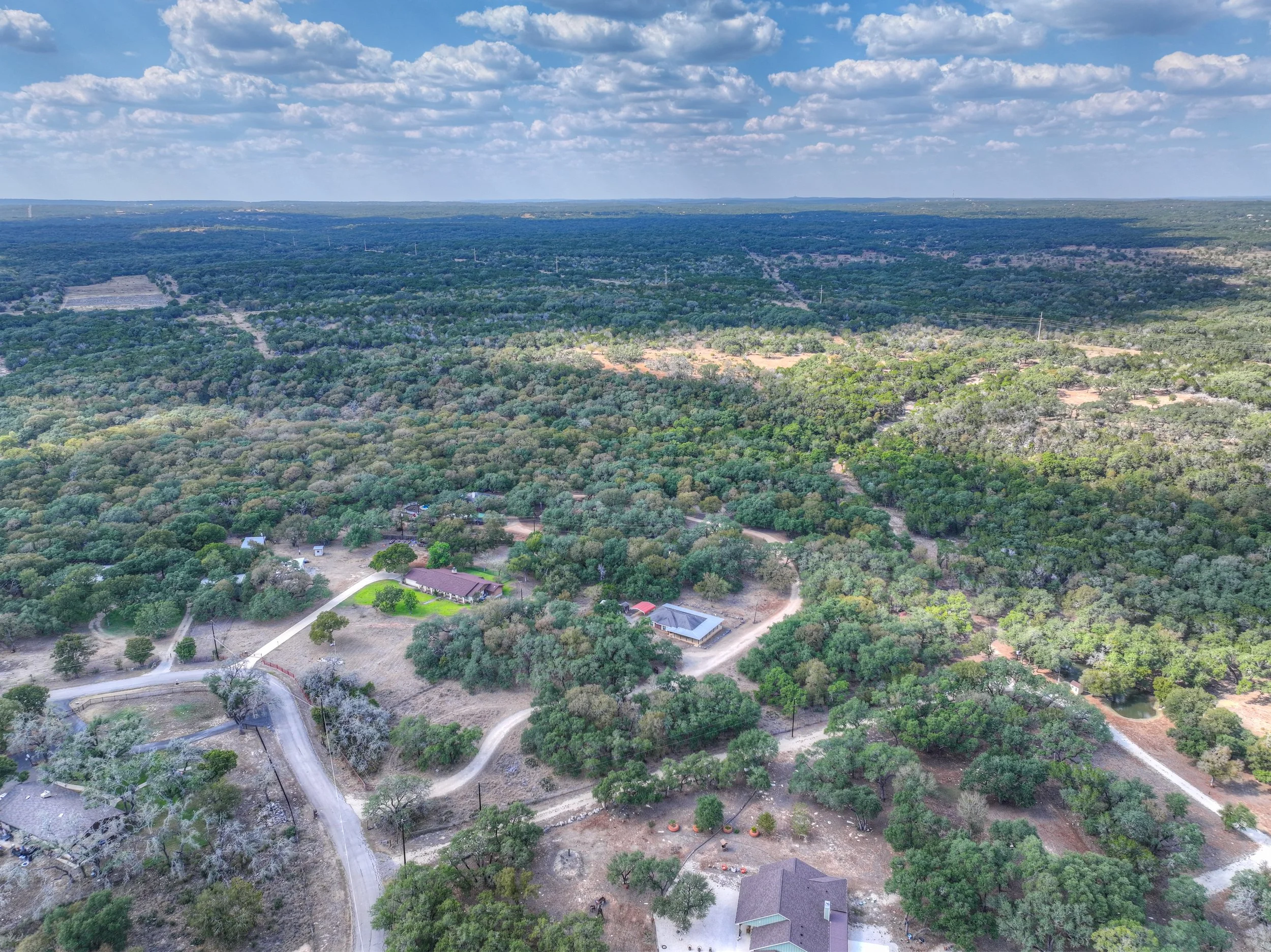 Aerial view of a rural landscape with dense trees, some open patches, winding roads, and a few buildings, under a partly cloudy sky.