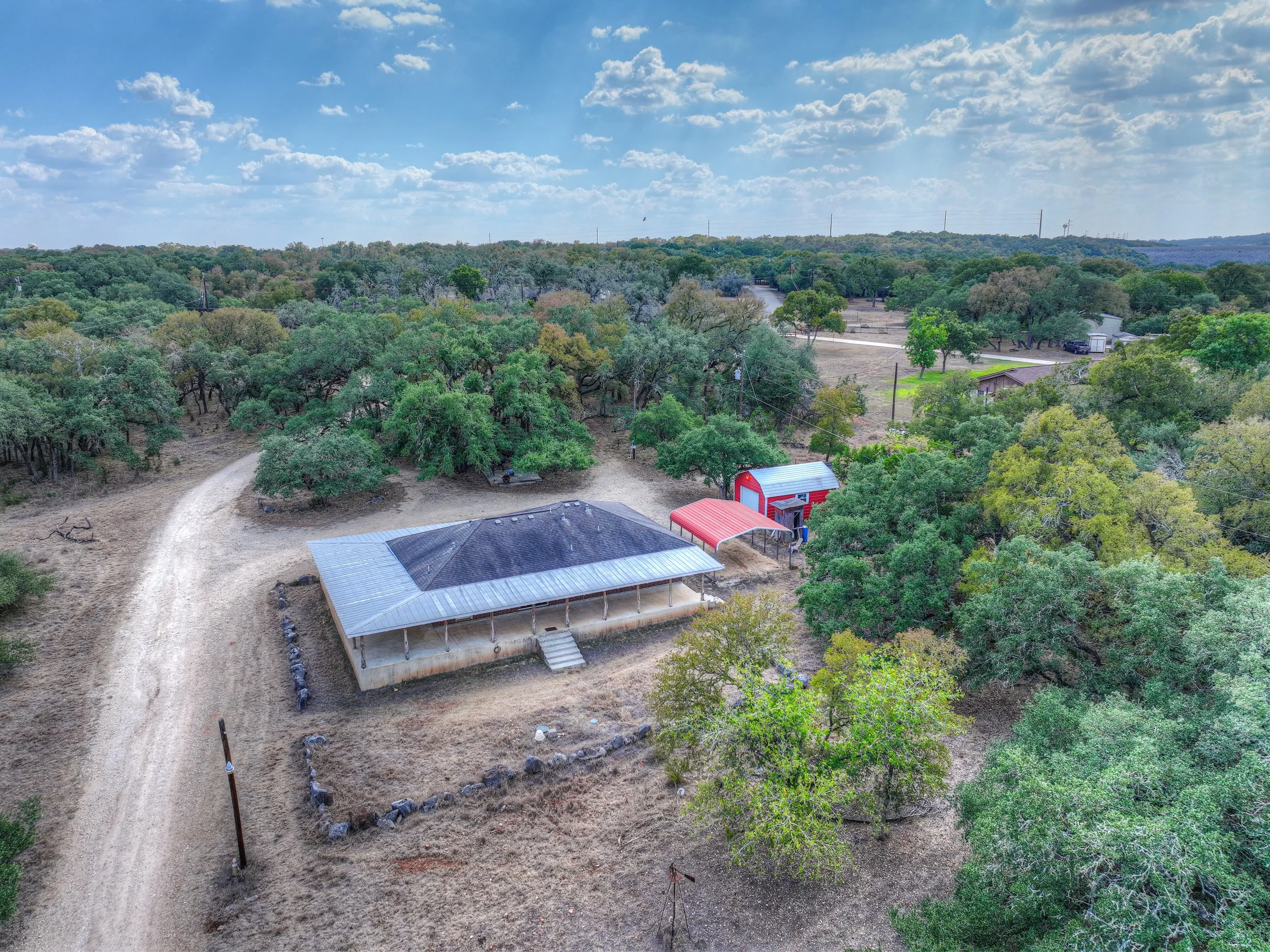 An aerial view of a rural property with a house featuring a porch, a dirt driveway, a red and white barn, a smaller red shed, and numerous trees and open fields under a partly cloudy sky.
