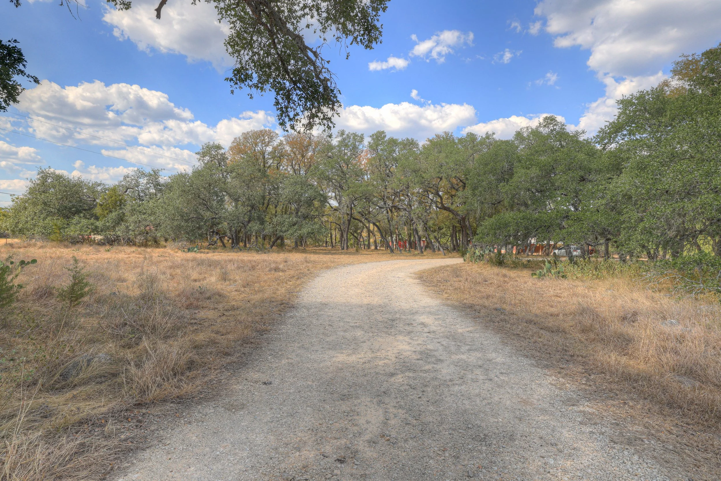 A dirt path winding through a dry grassy field with trees on either side under a partly cloudy sky.