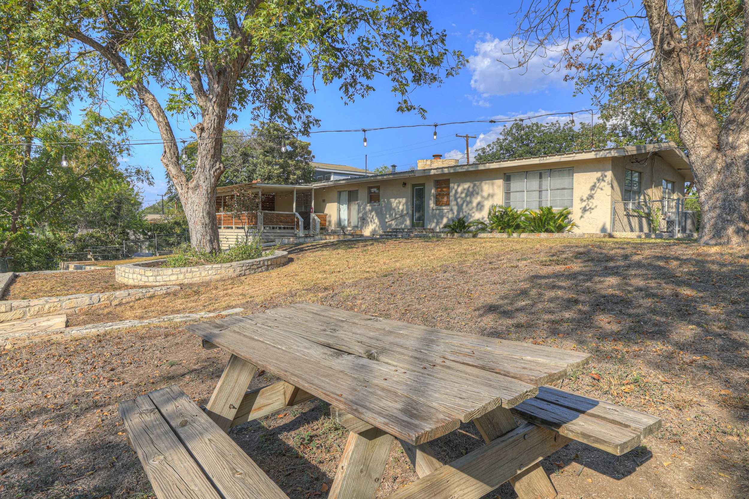 A backyard with a wooden picnic table on dirt ground, large trees providing shade, a house in the background with a small porch, windows, and a garden bed with plants, during daytime with a partly cloudy sky.