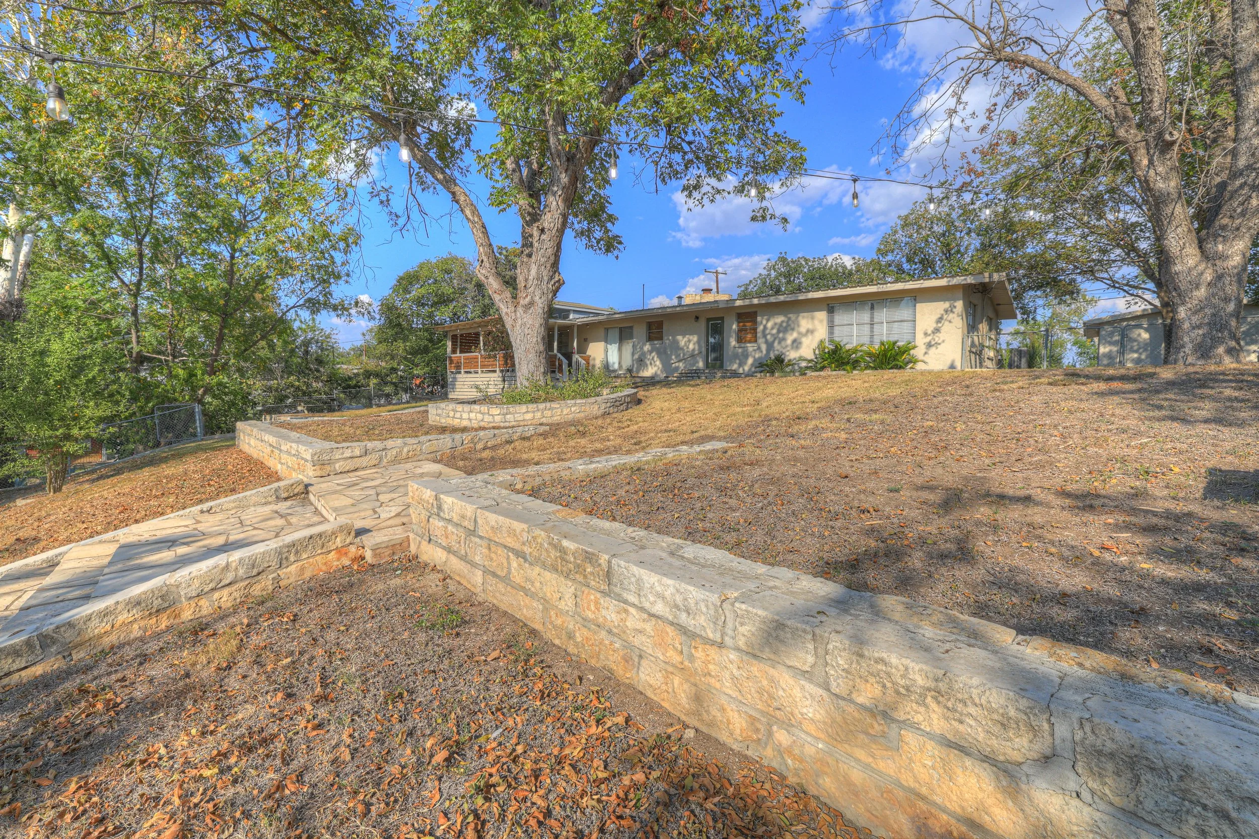 The backyard of a house with a stone pathway, large trees, and a clear blue sky.