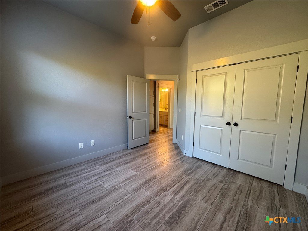 Empty bedroom with light gray walls, wooden flooring, ceiling fan, an open door leading to a bathroom, and a closet with double doors.