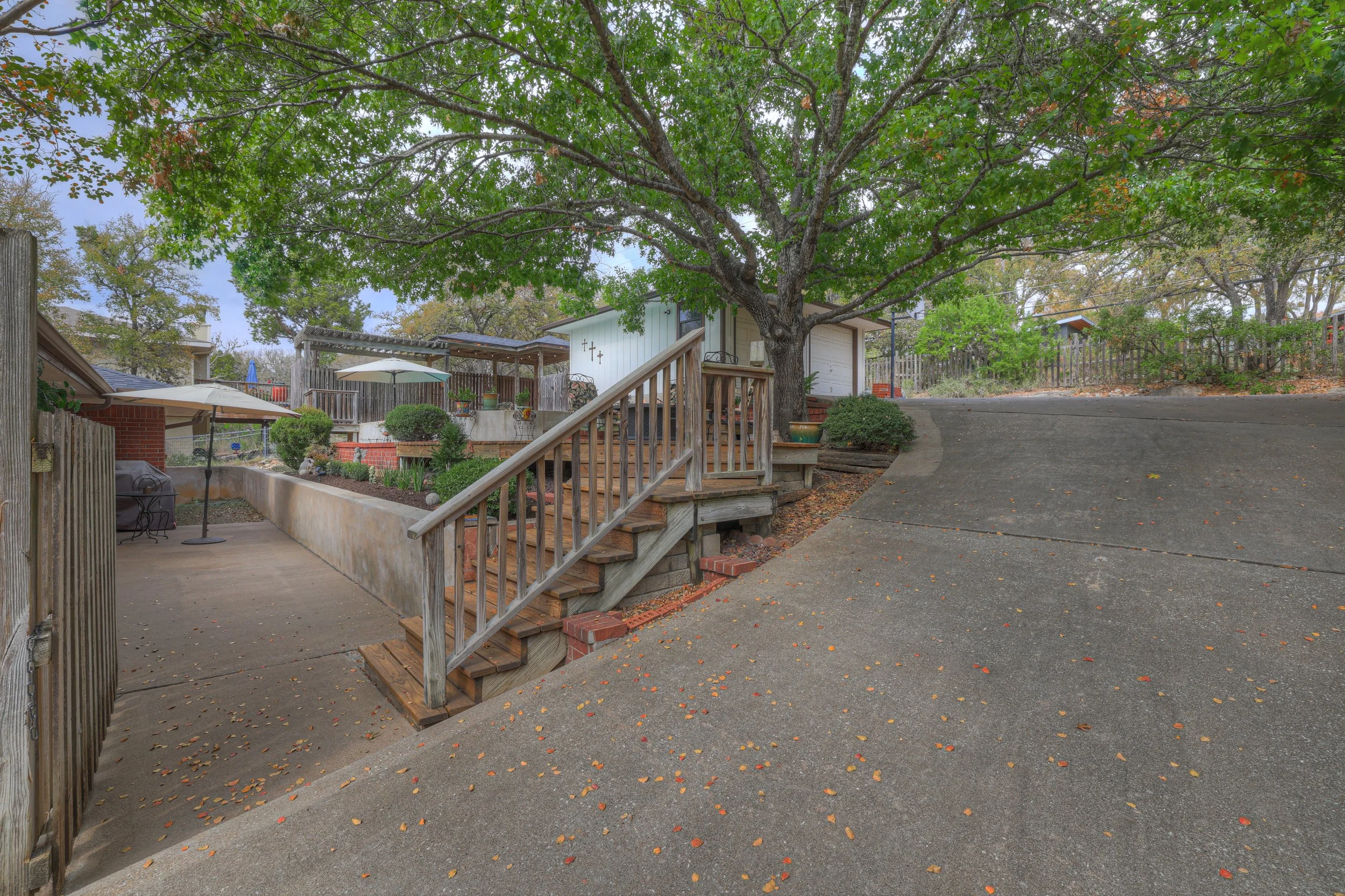 Residential backyard with a large tree, wooden stairs, a patio area with umbrellas, and a driveway.