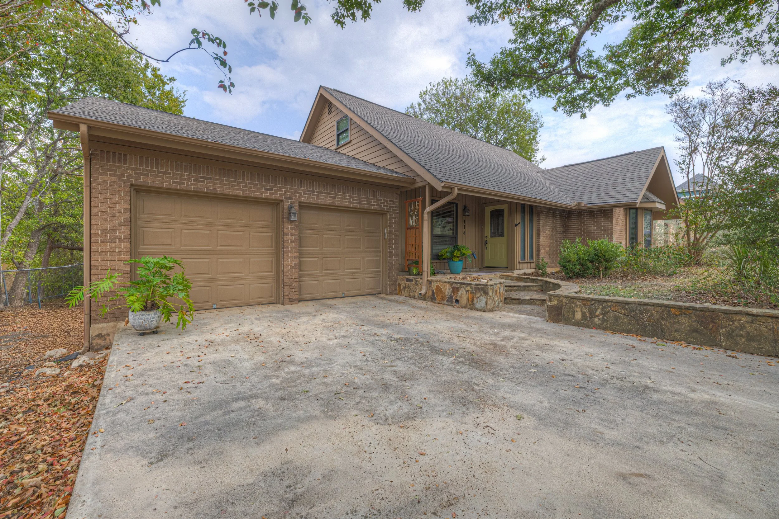 Front view of a brick house with a two-car garage, set in a garden with trees and plants, and a paved driveway leading to the garage.