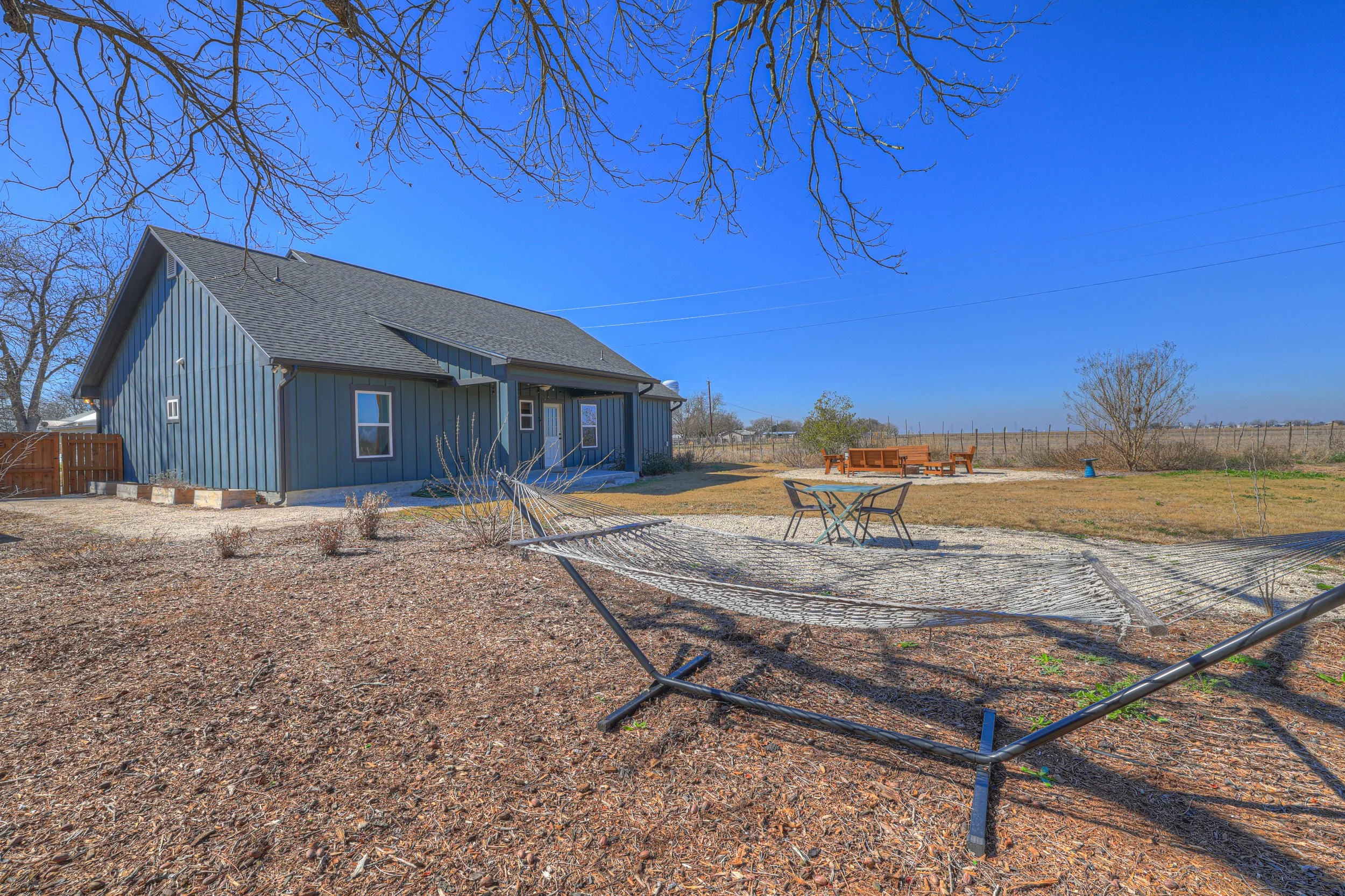 Backyard with a large house, a hammock, a table with chairs, a firepit, and benches, with dry grass and clear blue sky.