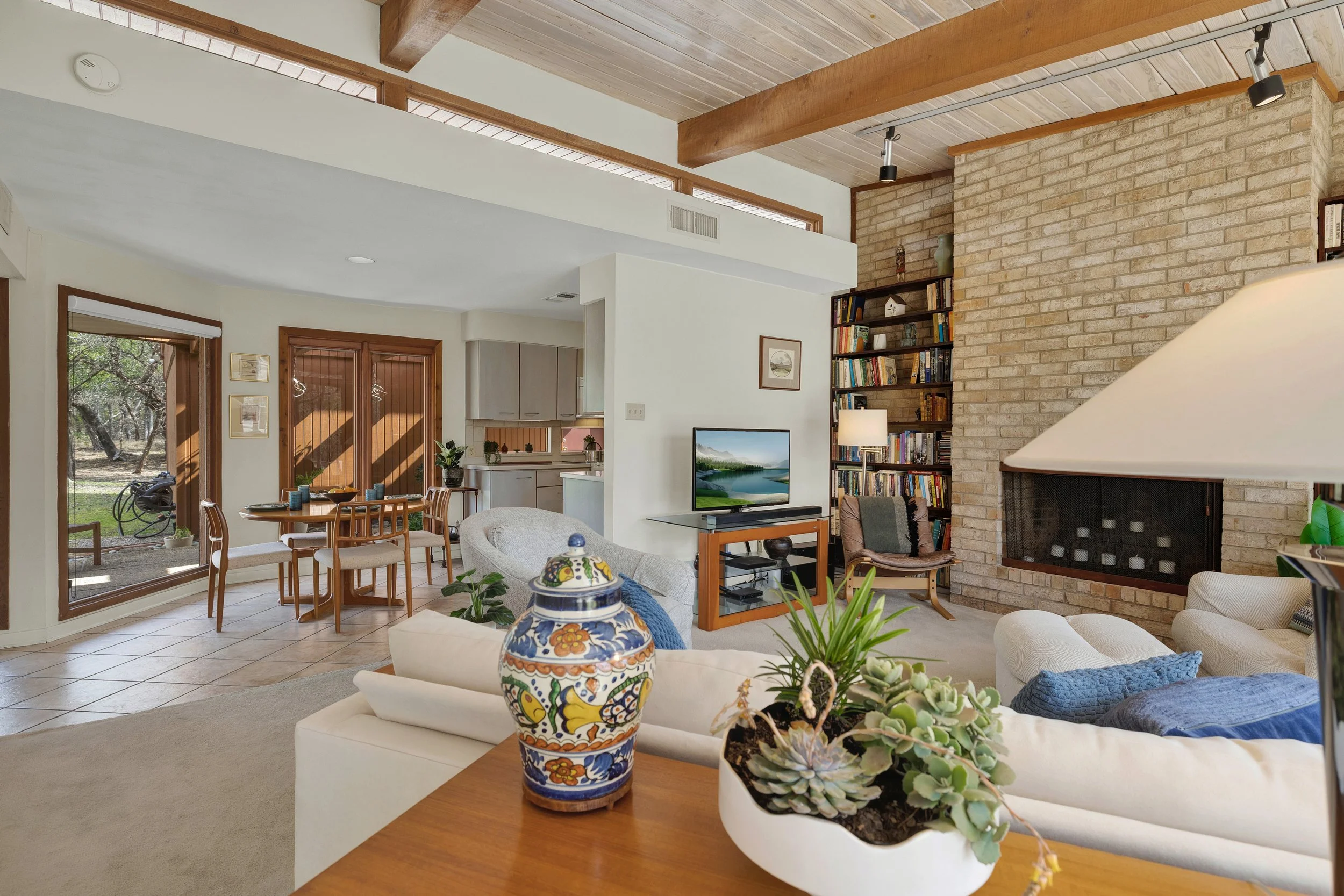 A cozy living room with a white sofa, a fireplace, a bookshelf, and a television. There is a dining area with a wooden table and chairs, and large windows and sliding glass doors showing an outdoor view.