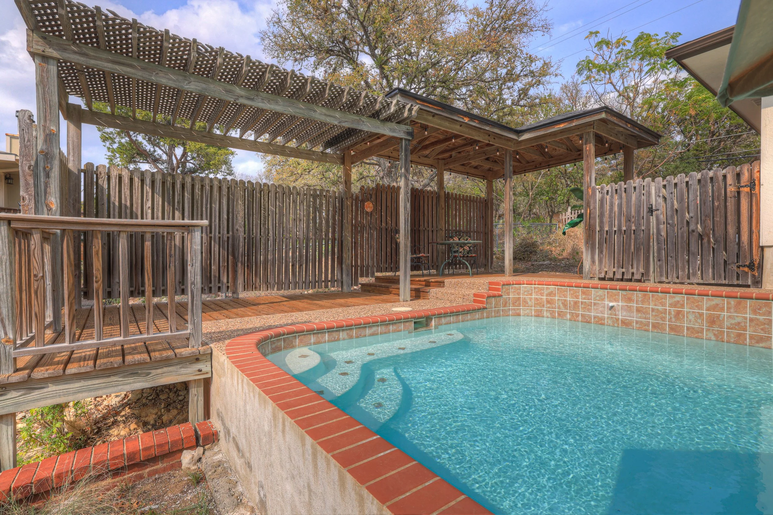 Backyard swimming pool with a brick edge, fencing, and a covered seating area with wooden posts.