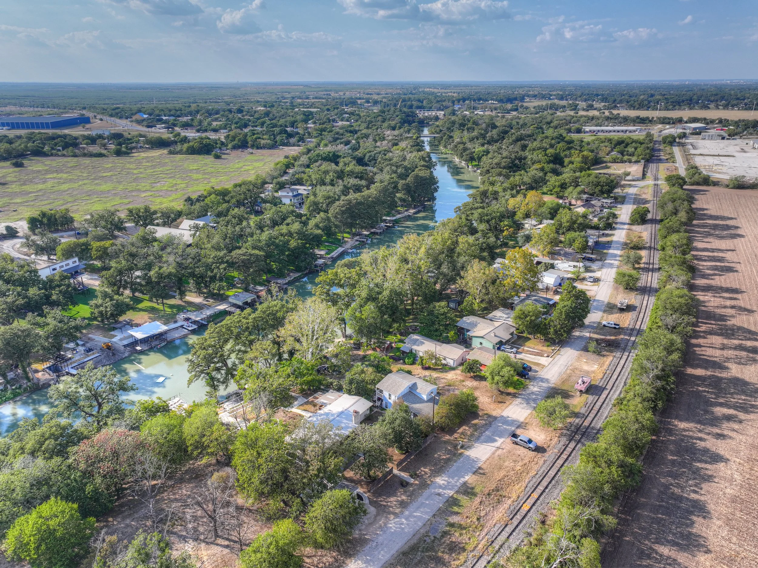 Aerial view of a residential canal neighborhood with houses along the water, trees, and a railroad track on the right side.