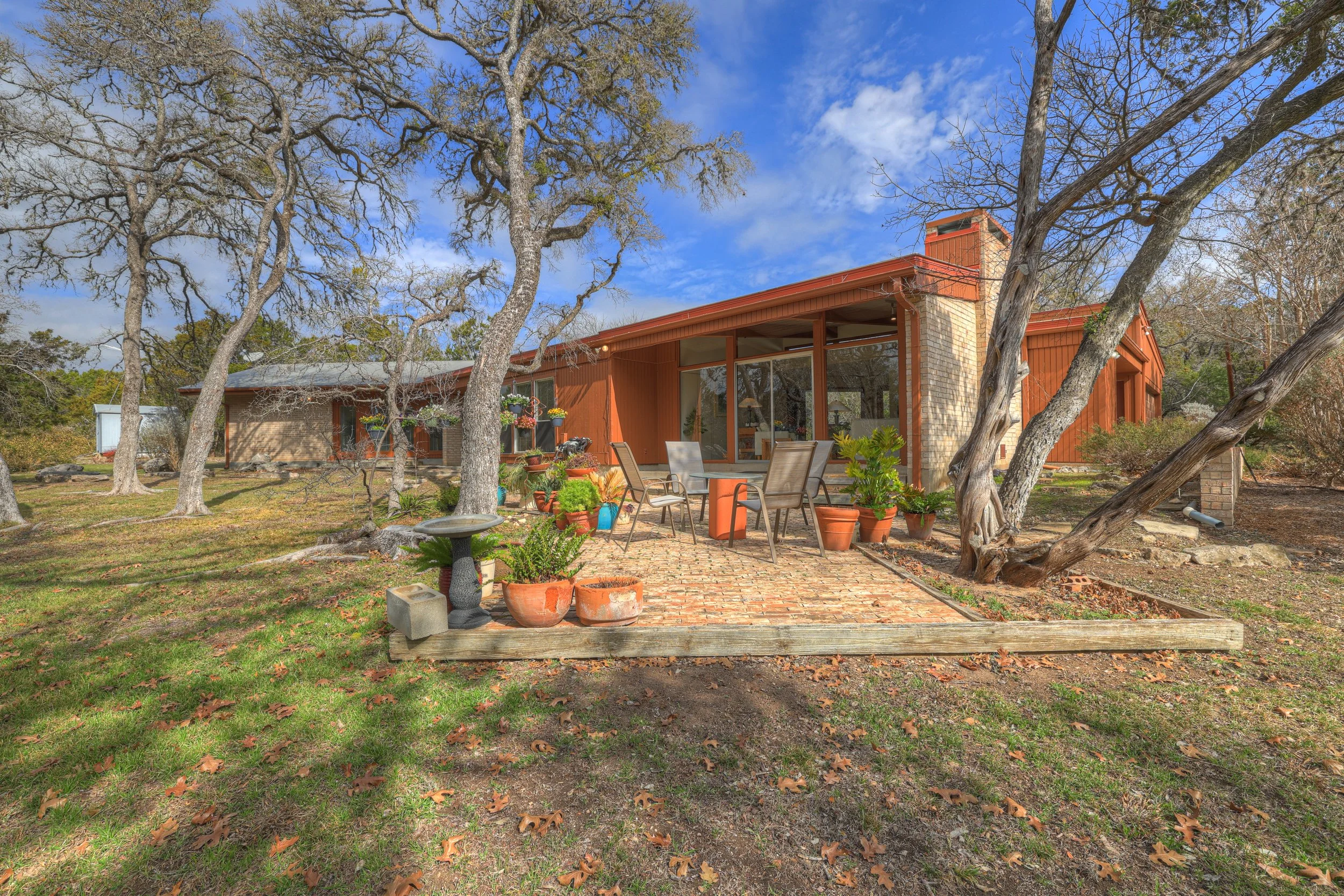 A mid-century modern house with a brick and wood exterior, large glass windows, and outdoor patio with chairs and potted plants, surrounded by leafless trees and a lawn, under a partly cloudy sky.