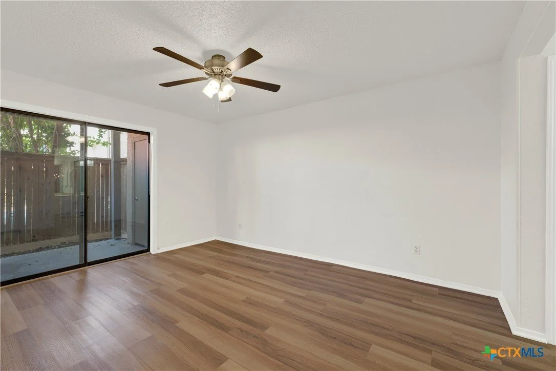 Empty living room with white walls, hardwood flooring, a ceiling fan with lights, and sliding glass door leading to a backyard patio.