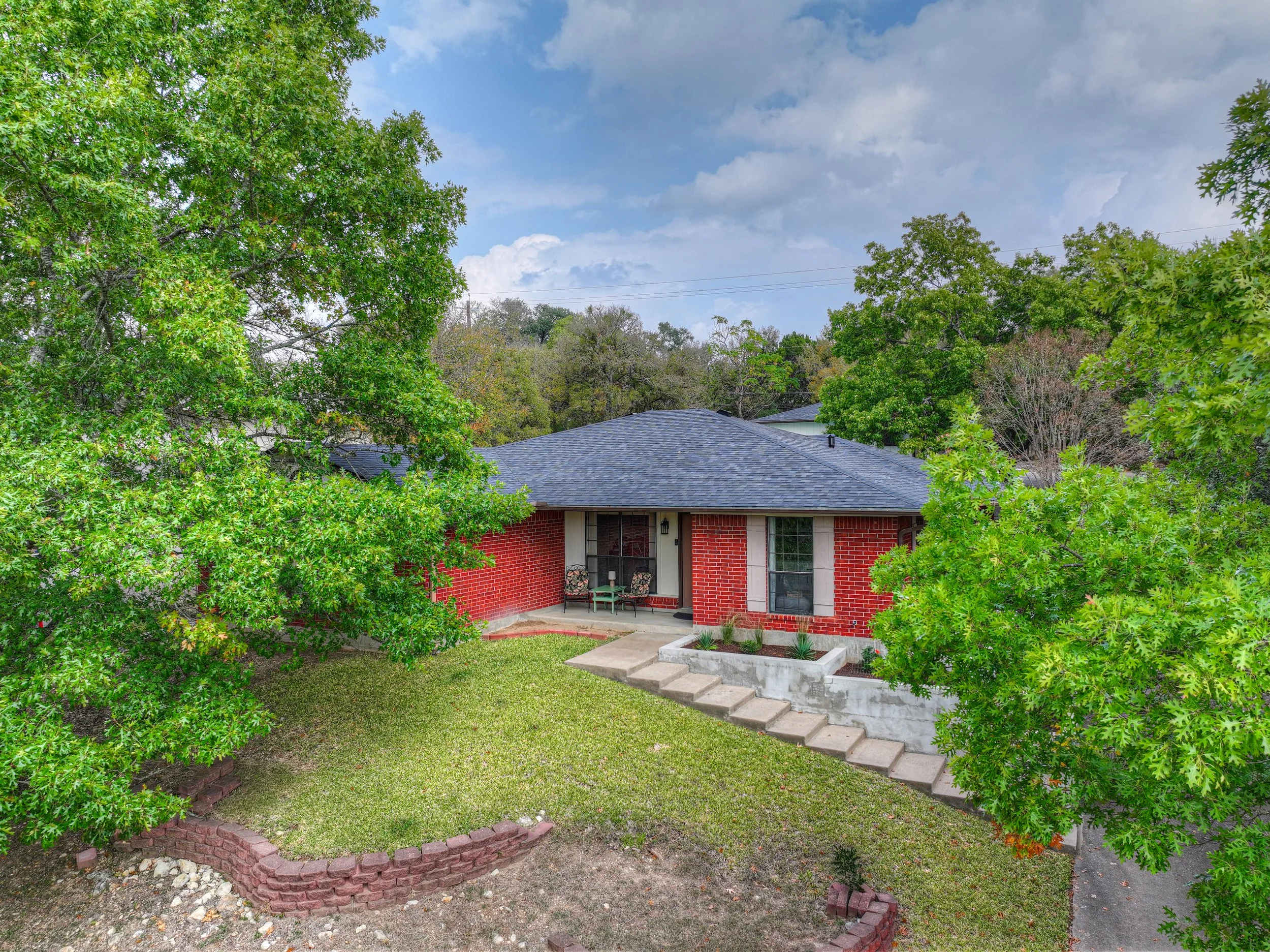 Front view of a single-story brick house with a dark gray shingle roof, surrounded by green trees and a lawn, with a small concrete porch and steps leading to the front door.